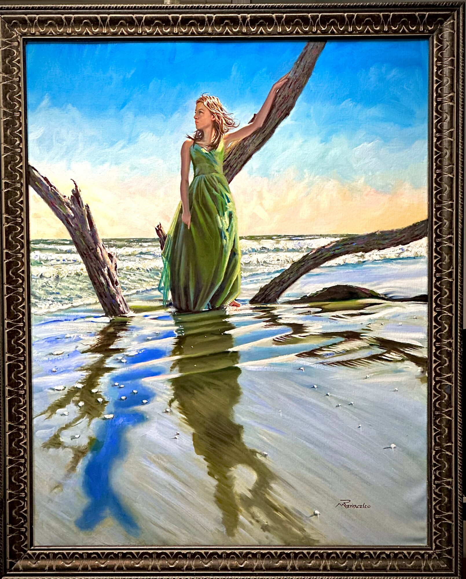 Woman in a green dress standing on a fallen tree in water, with a blue sky and ocean in background.