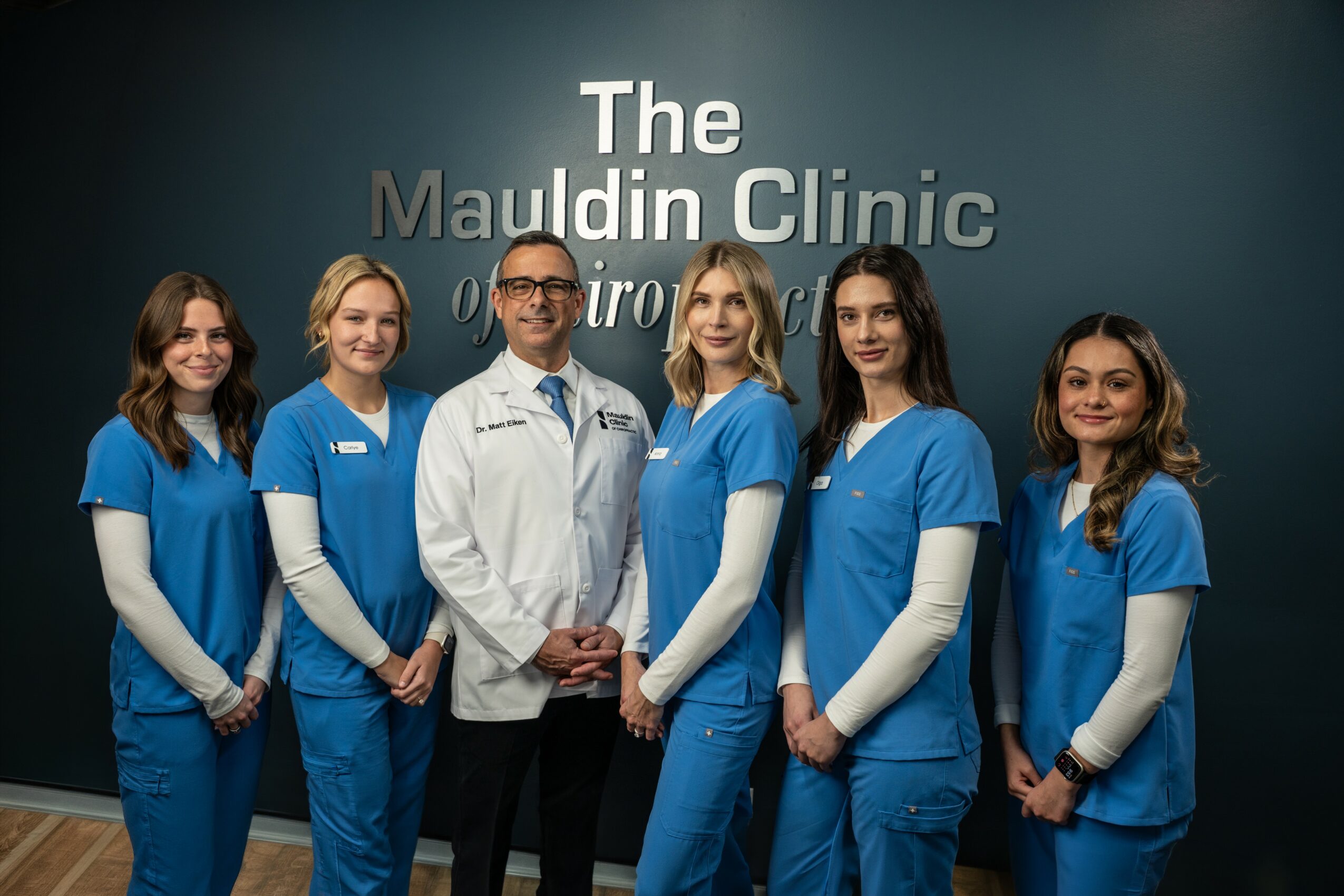 Six medical professionals in blue scrubs and white lab coat standing in front of a wall with the clinic's name.