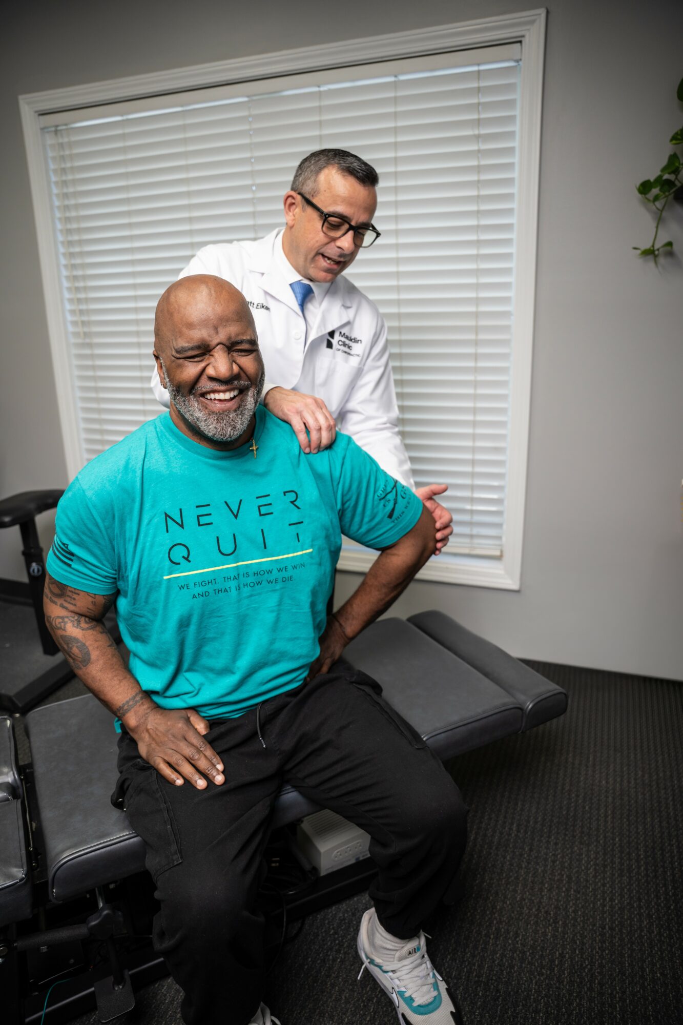 Two men in a medical office, one sitting and smiling, the other standing behind, smiling and touching the seated man's shoulder.