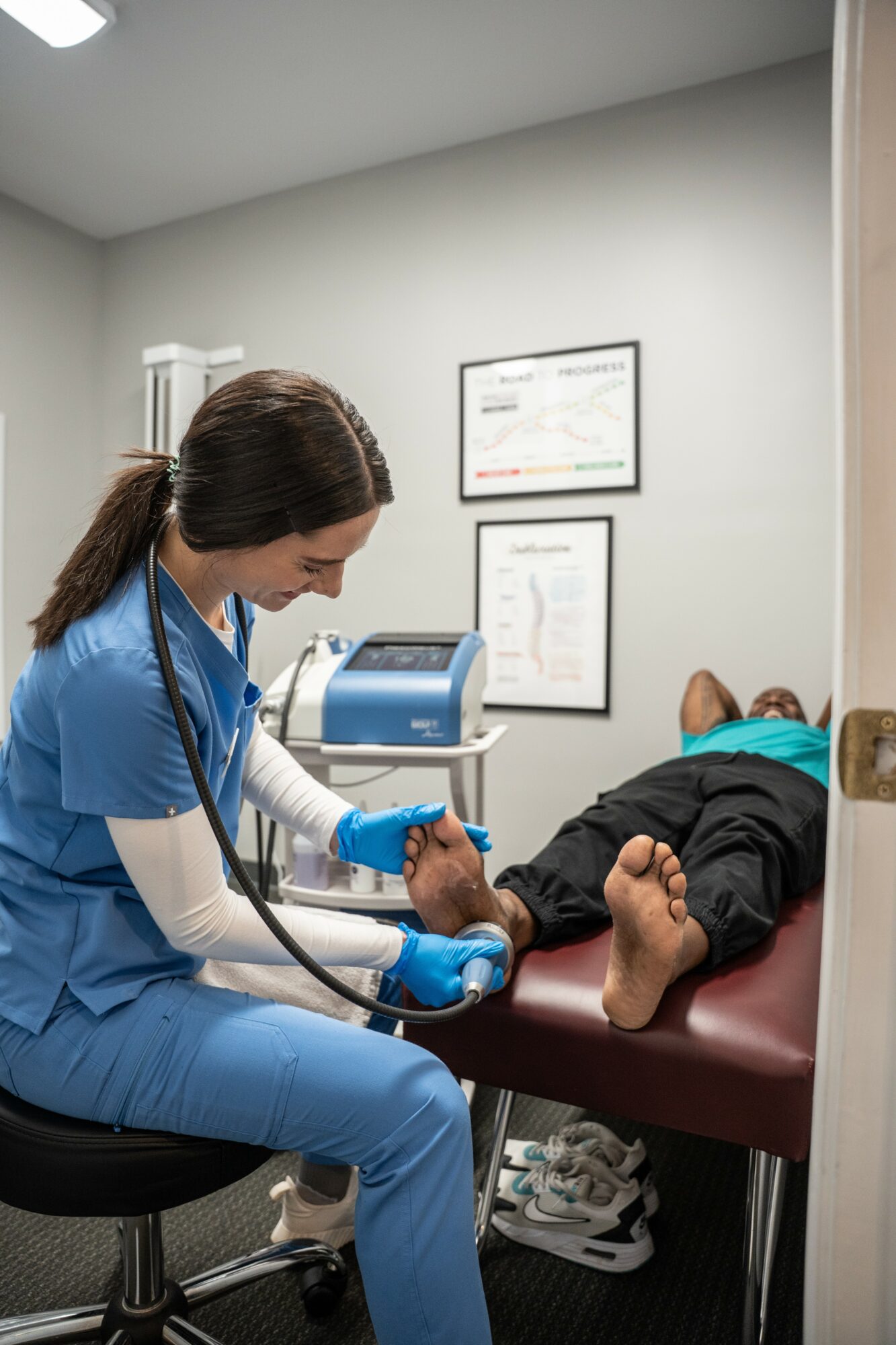 Healthcare professional in blue scrubs using medical equipment on a patient lying on an examination table in a clinical room.