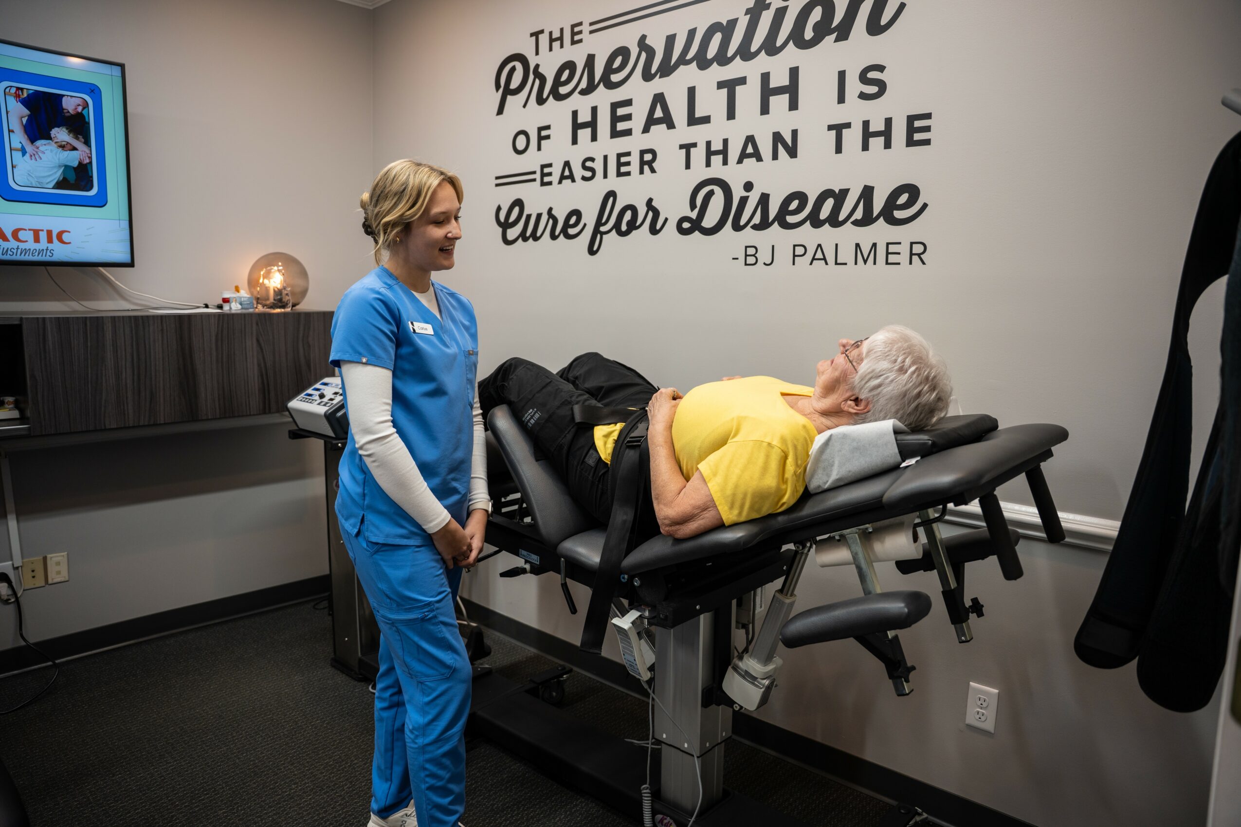 Healthcare professional in blue scrubs standing beside an elderly woman lying on a medical examination table in a clinic room.