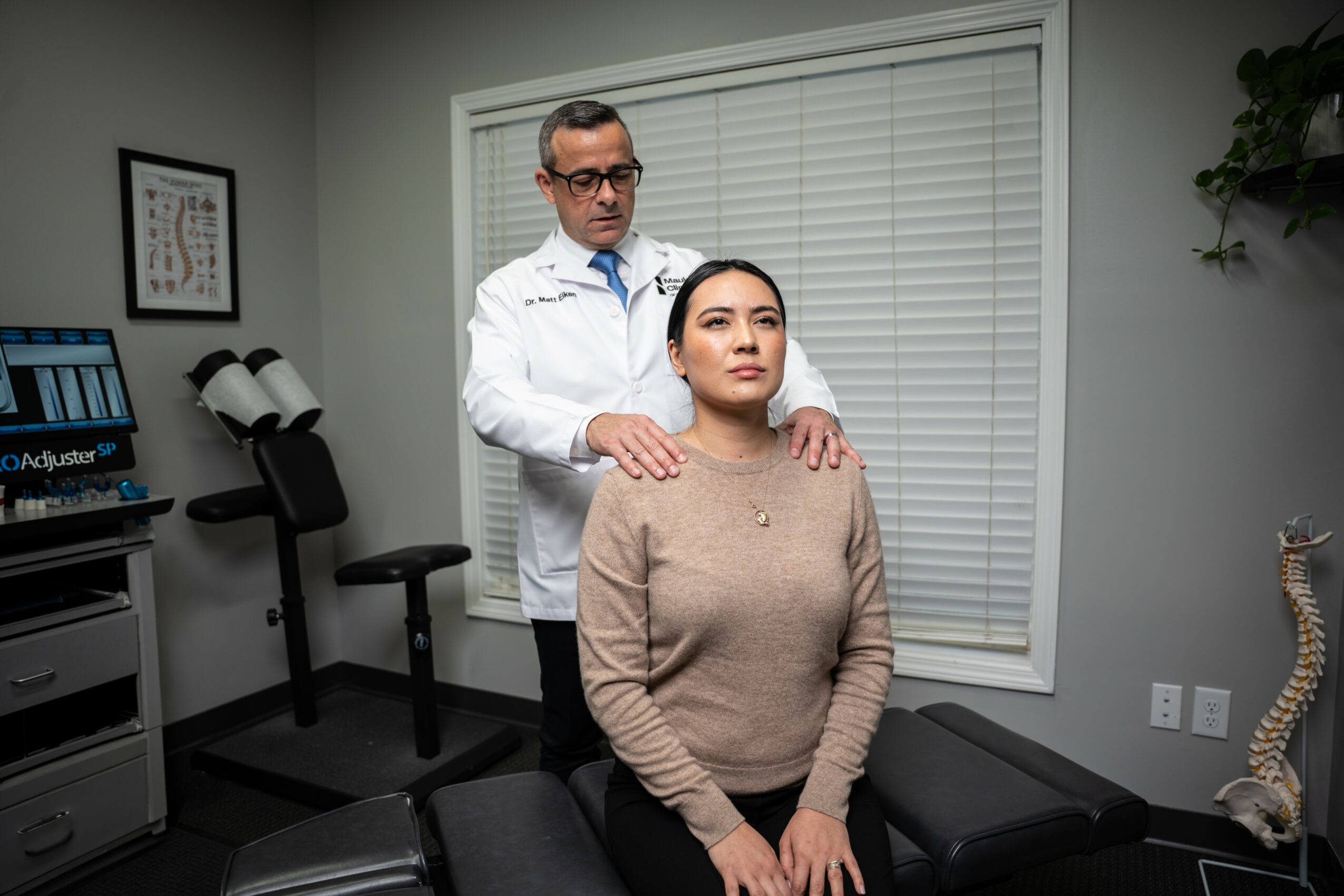 Doctor standing behind woman in medical office, hands on her shoulders, woman sitting with eyes closed, neutral expression.