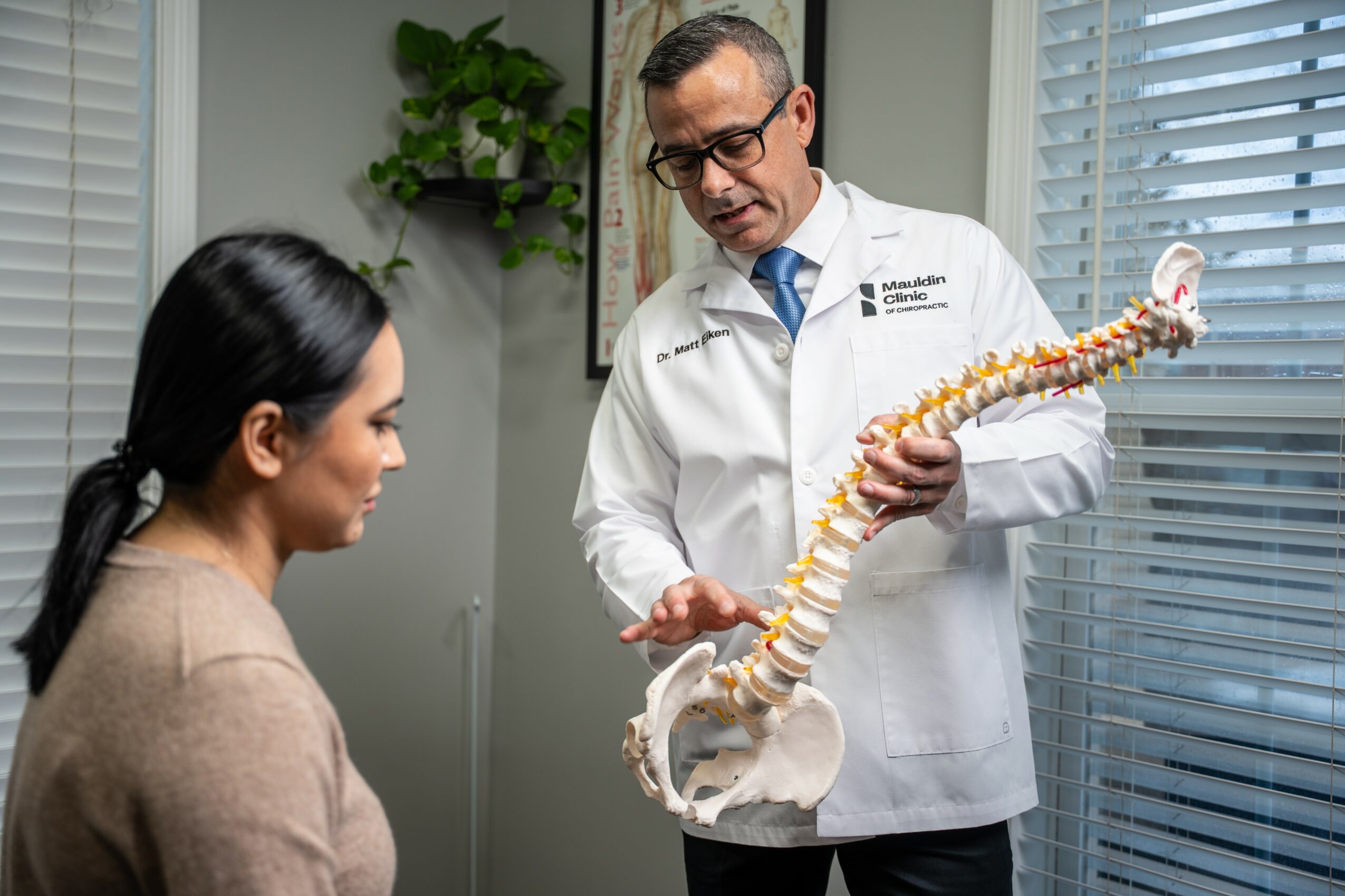 Doctor explains spinal model to a woman in a medical office with window blinds and a plant.