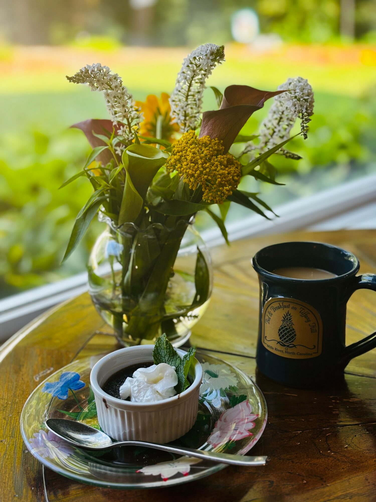 Vase with white and yellow flowers on a wooden table, a cup, and a spoon, with a window view of greenery outside.