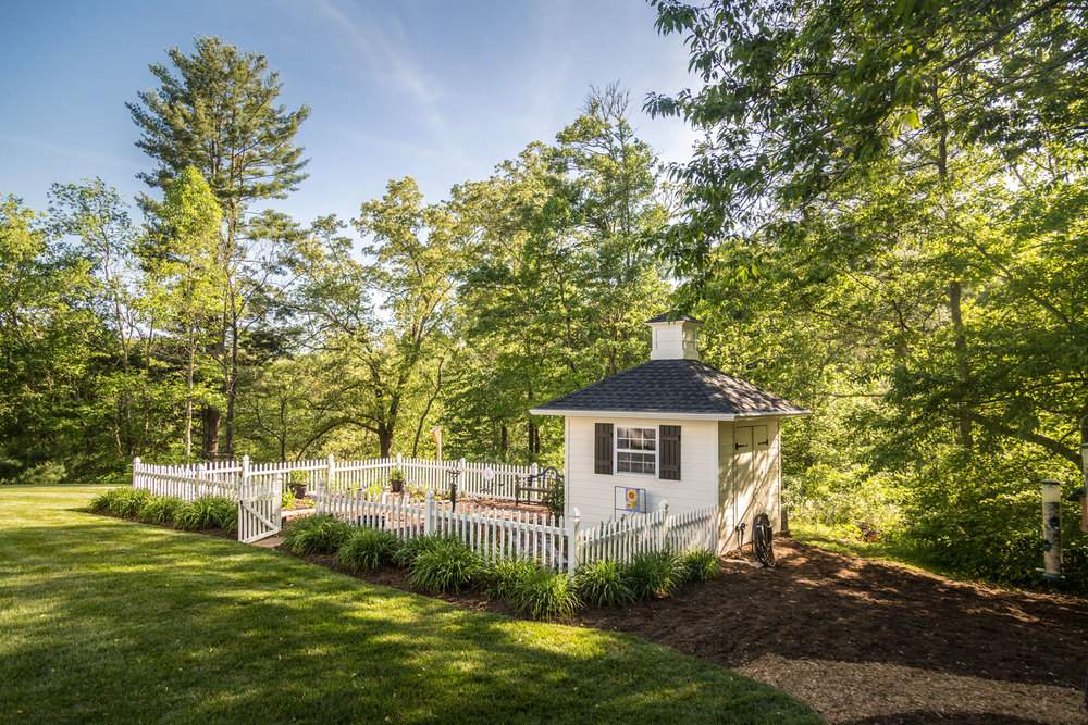 Small white house with a porch, surrounded by trees and a white fence, on a sunny day.