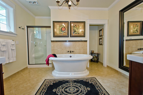 Bathroom with bathtub, shower, mirror, and framed artwork, beige walls, and a black rug with intricate pattern.