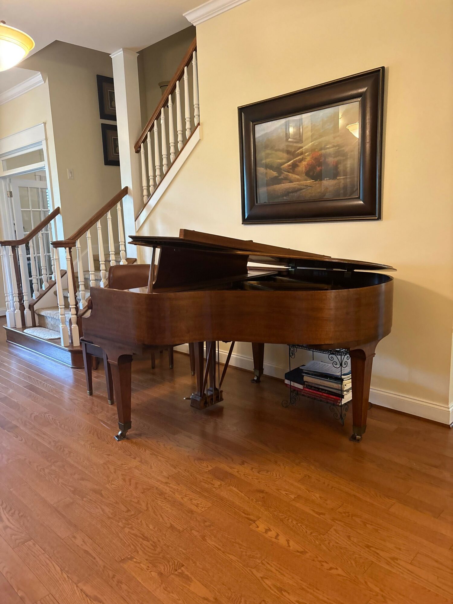Brown grand piano with open lid, placed against a beige wall with a framed landscape painting above, on a wooden floor.