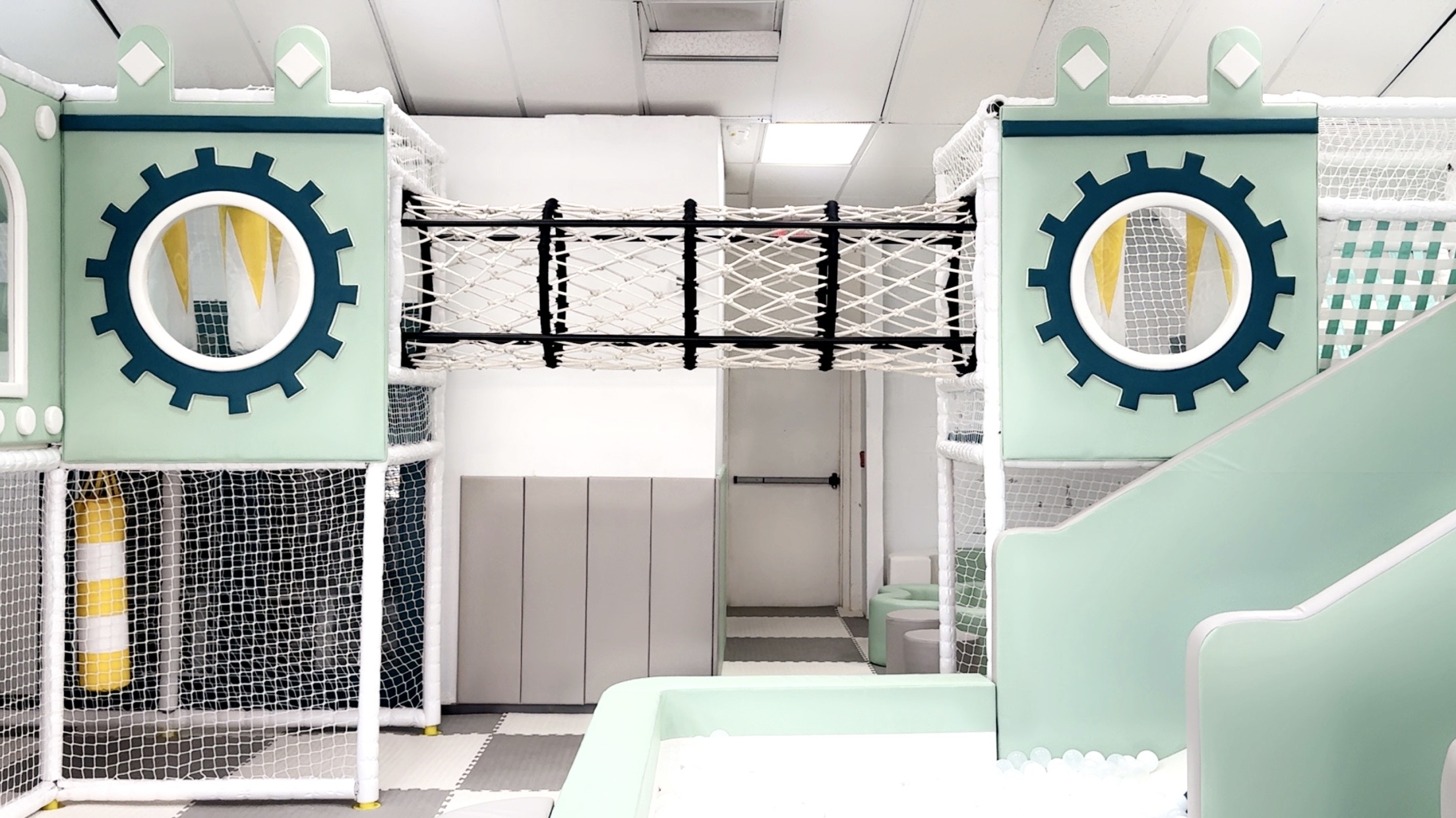 Indoor playground with gear-shaped structures and a bridge, featuring soft play equipment and a slide.