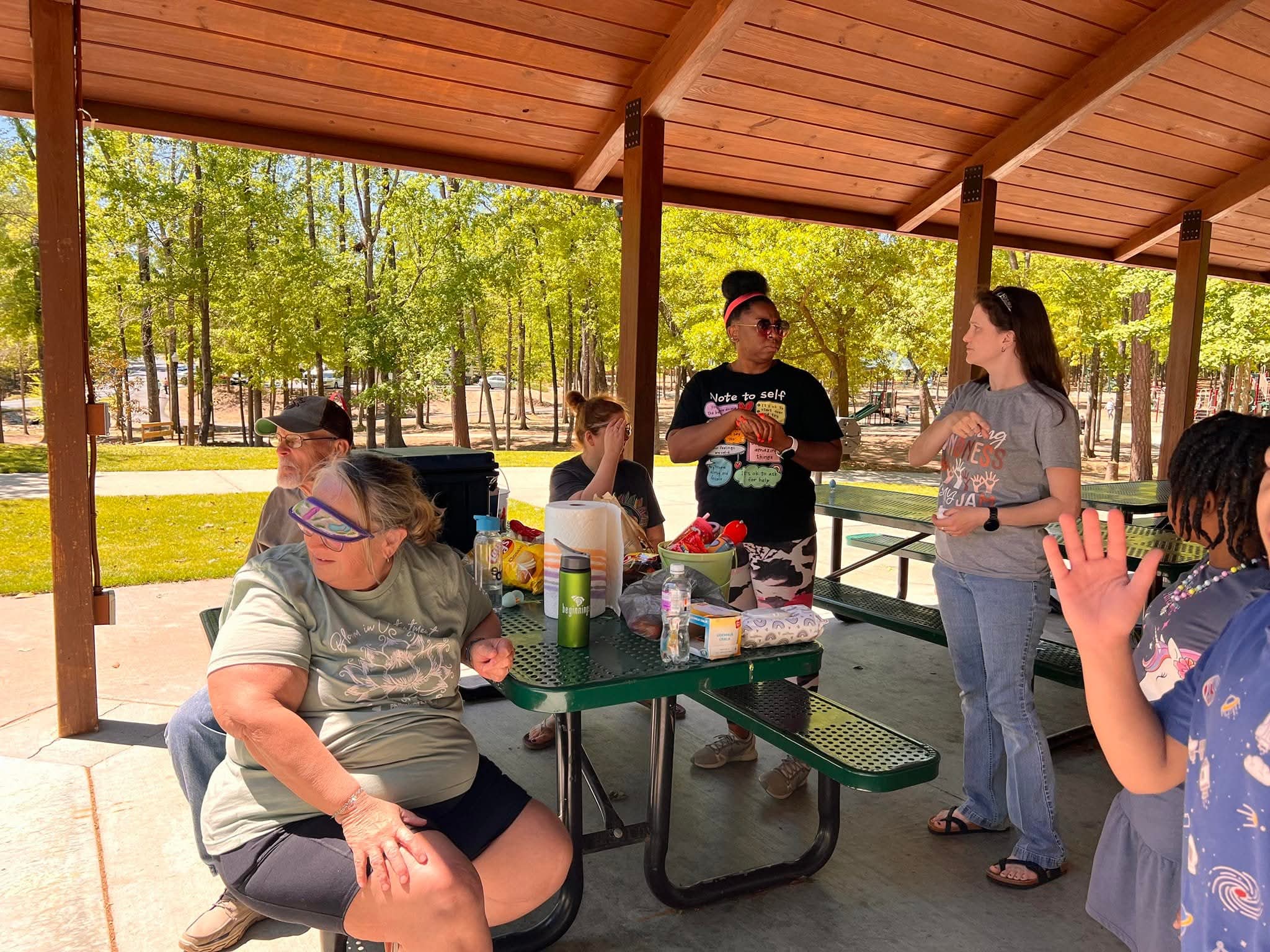 Group of people under a pavilion with trees and grass in the background, standing and talking around a picnic table.