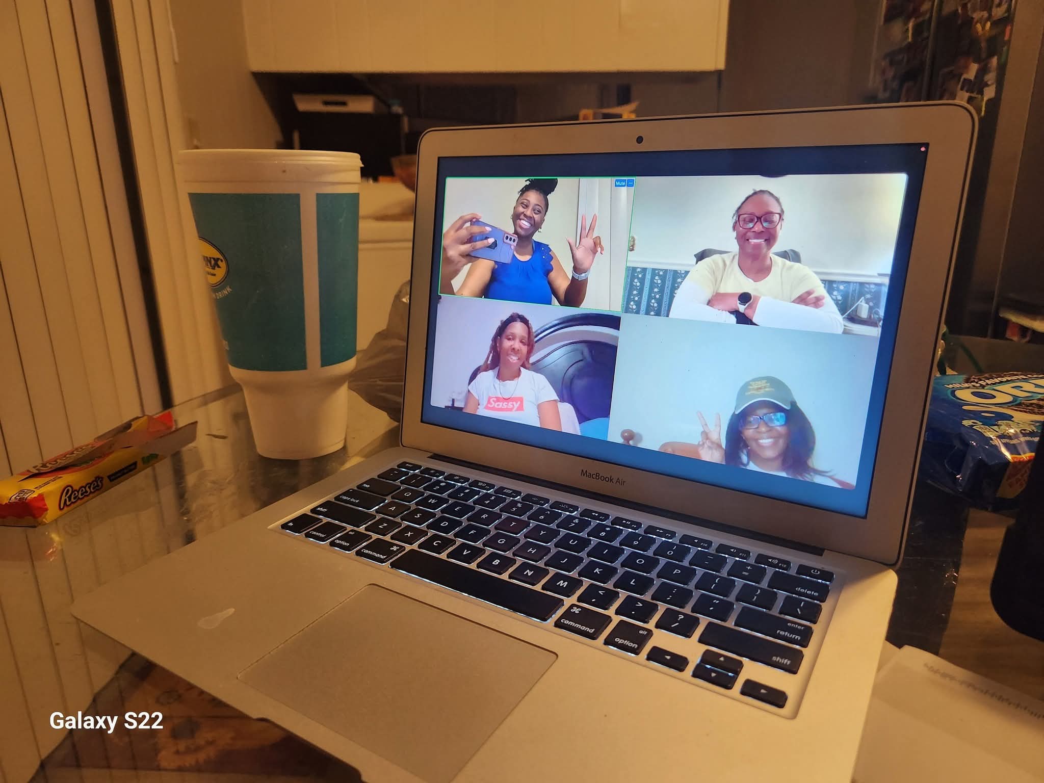 Laptop screen showing a video call with five people smiling and waving, in a room with various objects around.