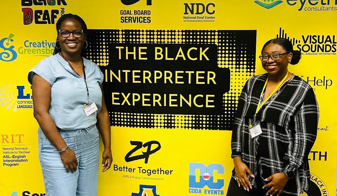 Two women standing in front of a yellow and black event backdrop with text and logos.