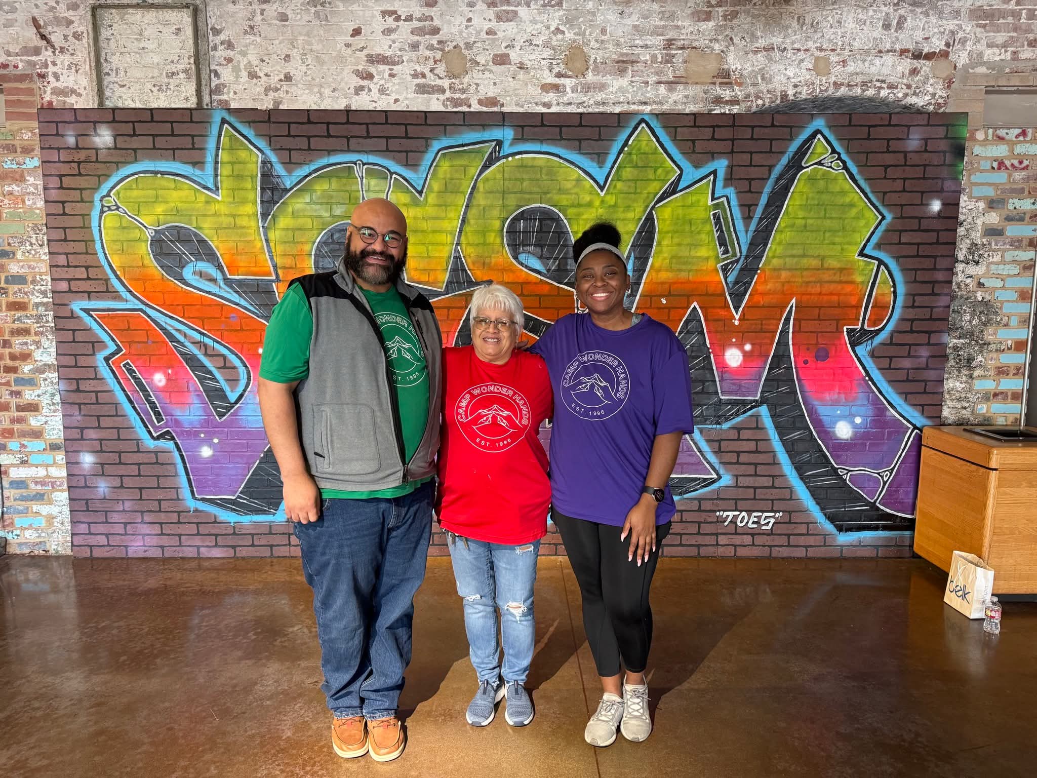 Three people stand in front of colorful graffiti on a brick wall, smiling at the camera.