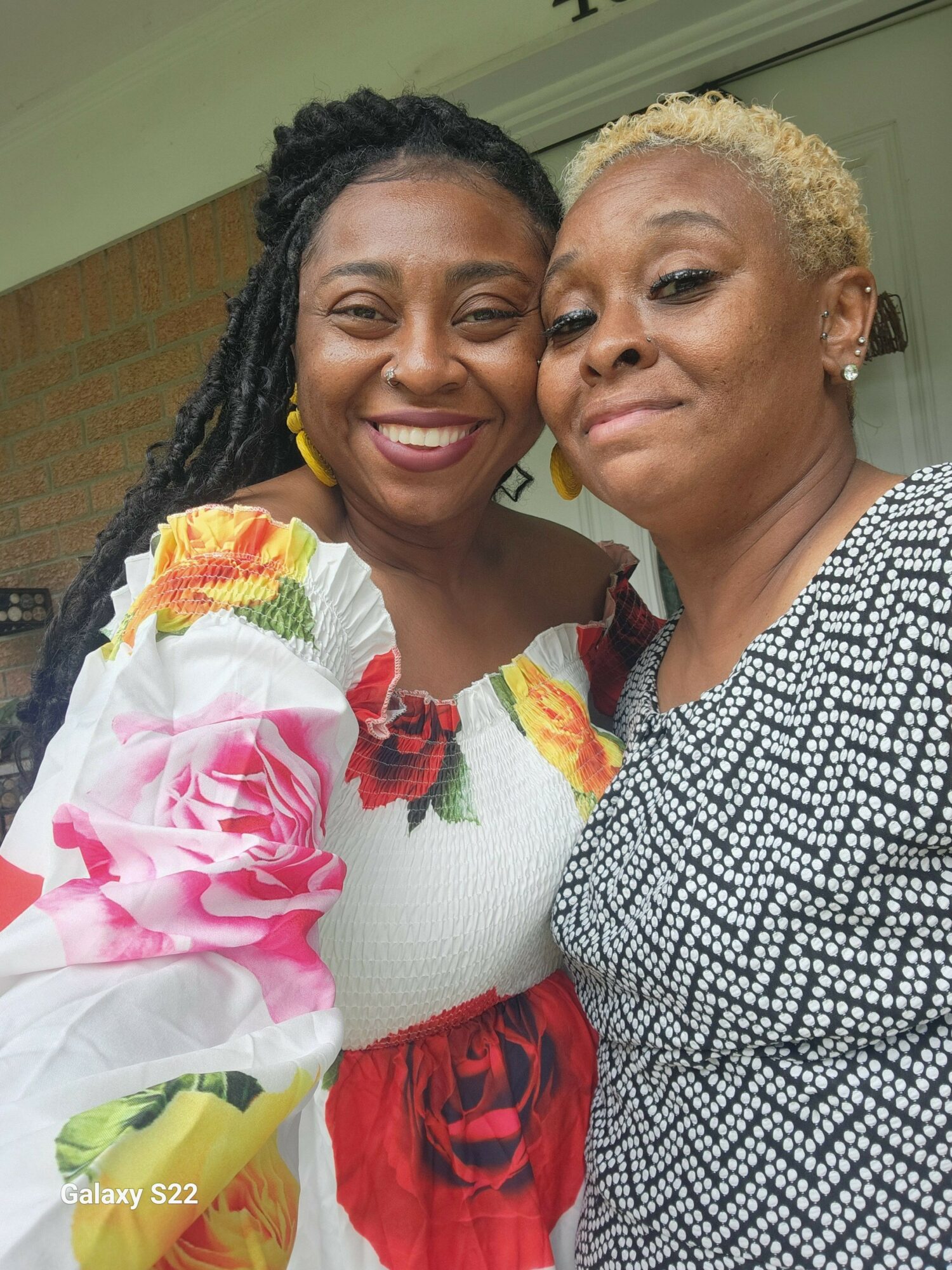Two women smiling closely, one with dark braided hair and colorful floral blouse, the other with short blonde hair and patterned top.