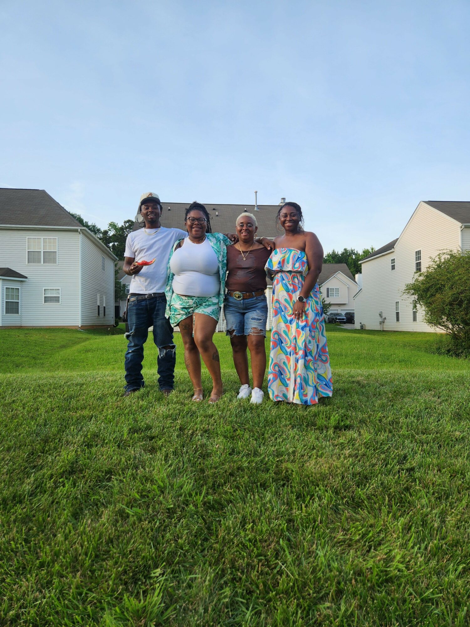 Four people standing on grass in front of houses, smiling, with clear sky above.