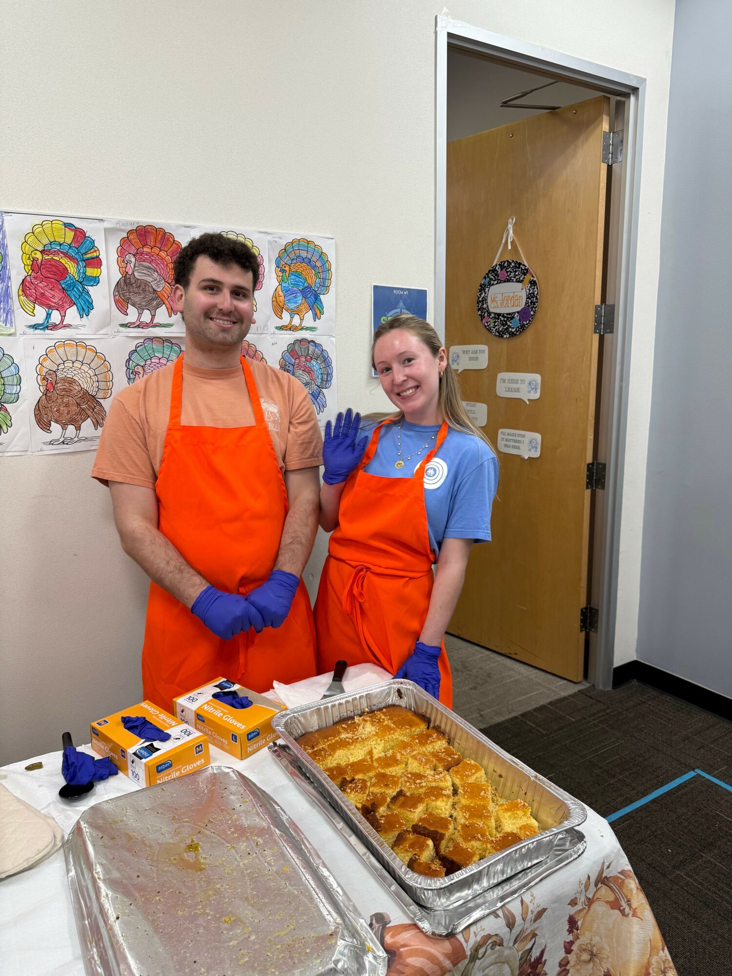 Two people in orange aprons and blue gloves standing behind a tray of food in a kitchen or cafeteria setting.