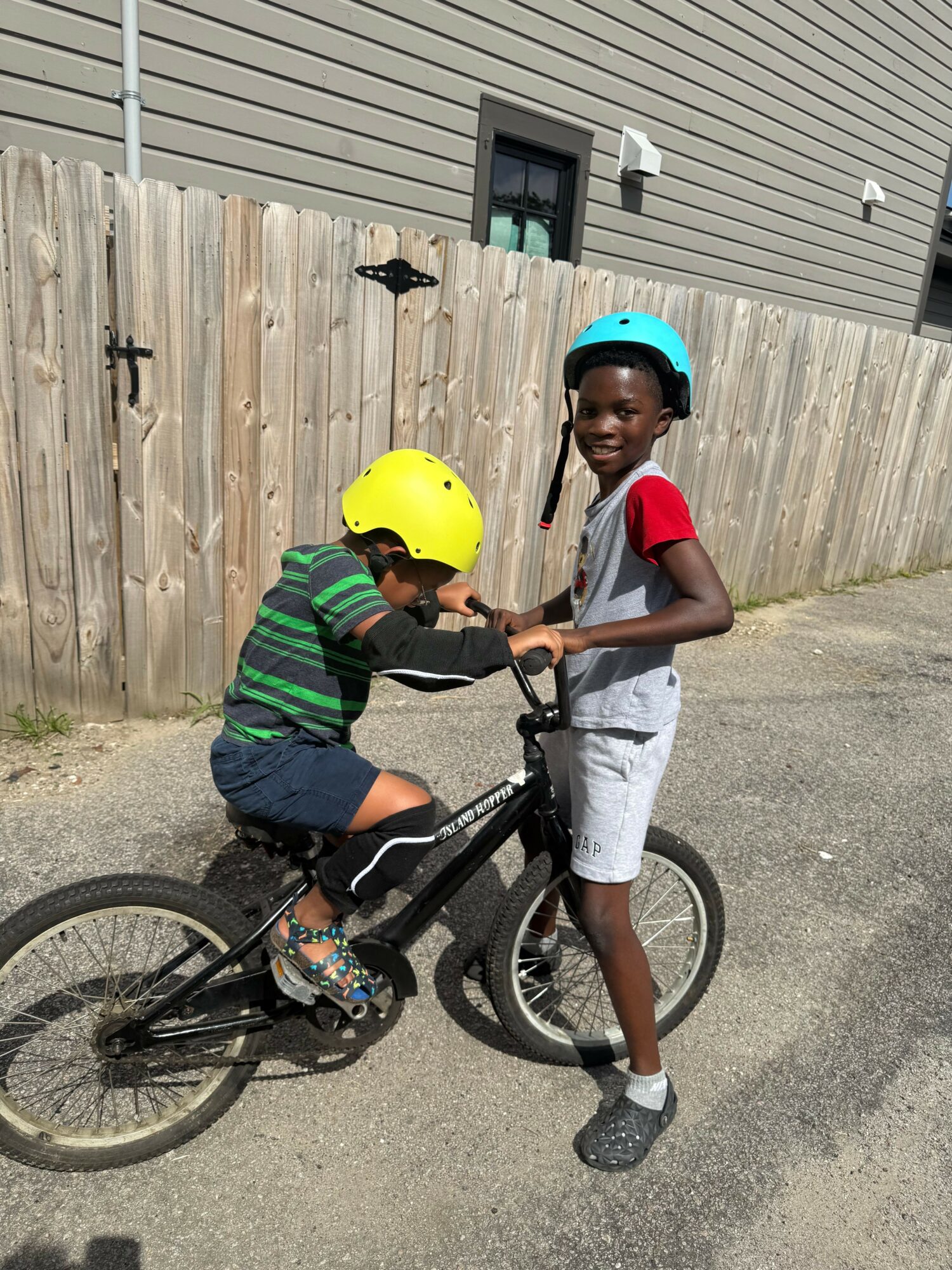Two children with helmets, one sitting on a bicycle and the other standing beside, smiling outdoors near a wooden fence.