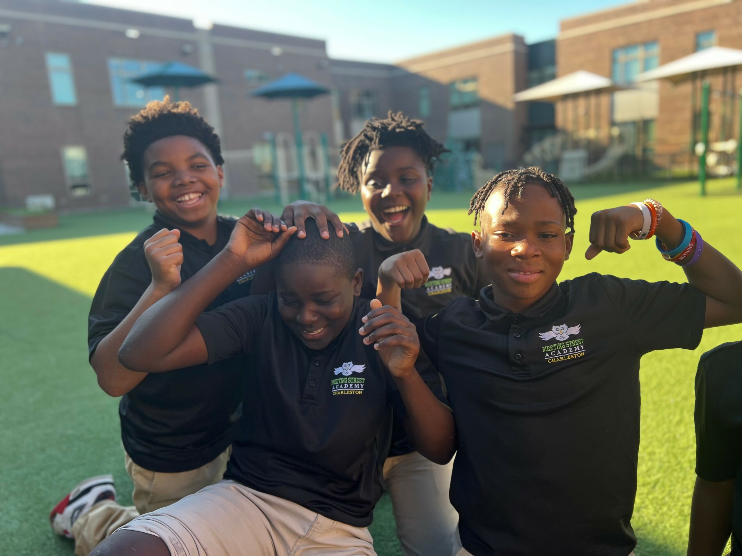 Four children smiling and flexing their arms outdoors on a sunny day.