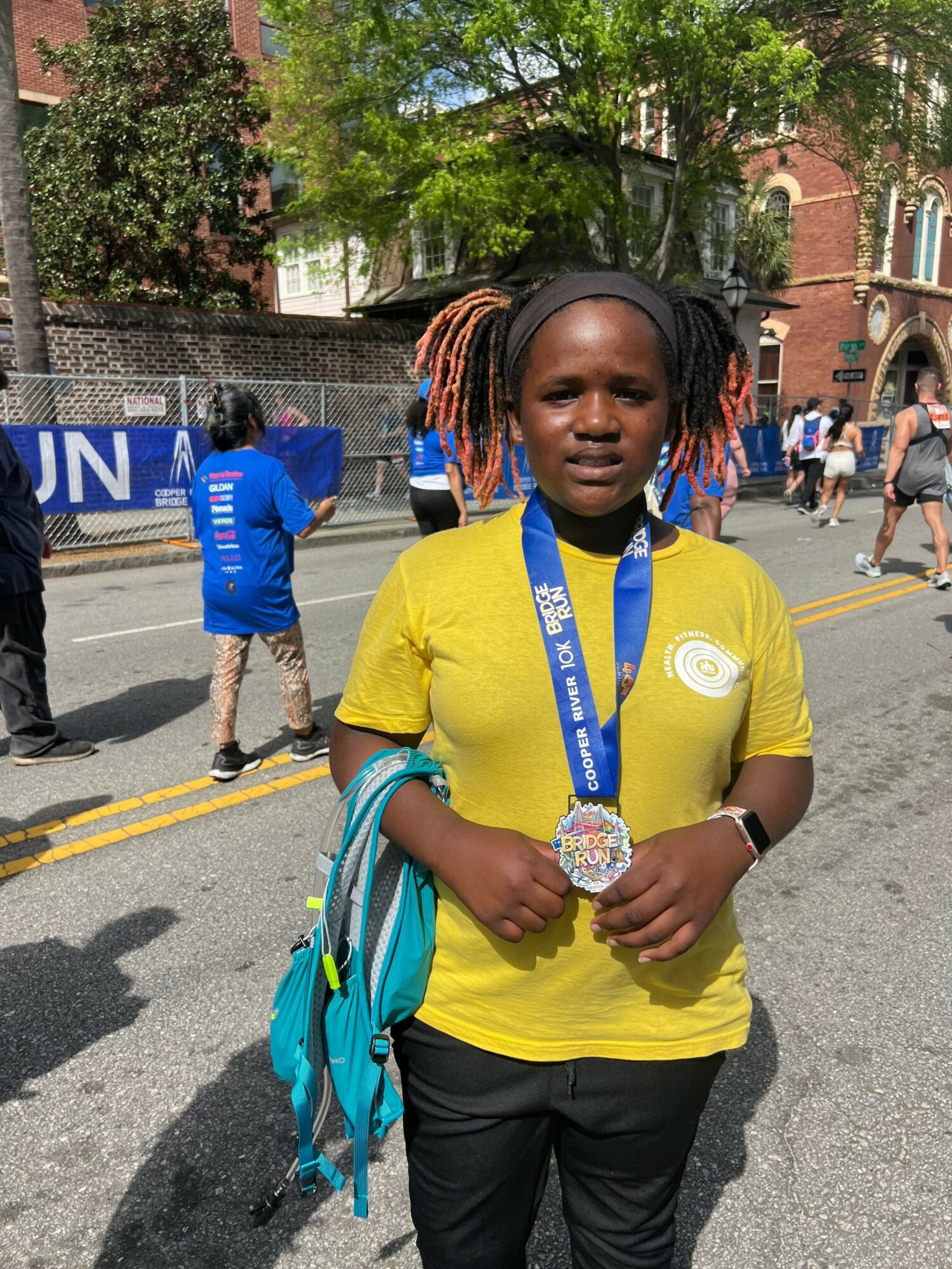 Young woman with braided hair wearing a yellow shirt and holding a medal, standing outdoors on a street.