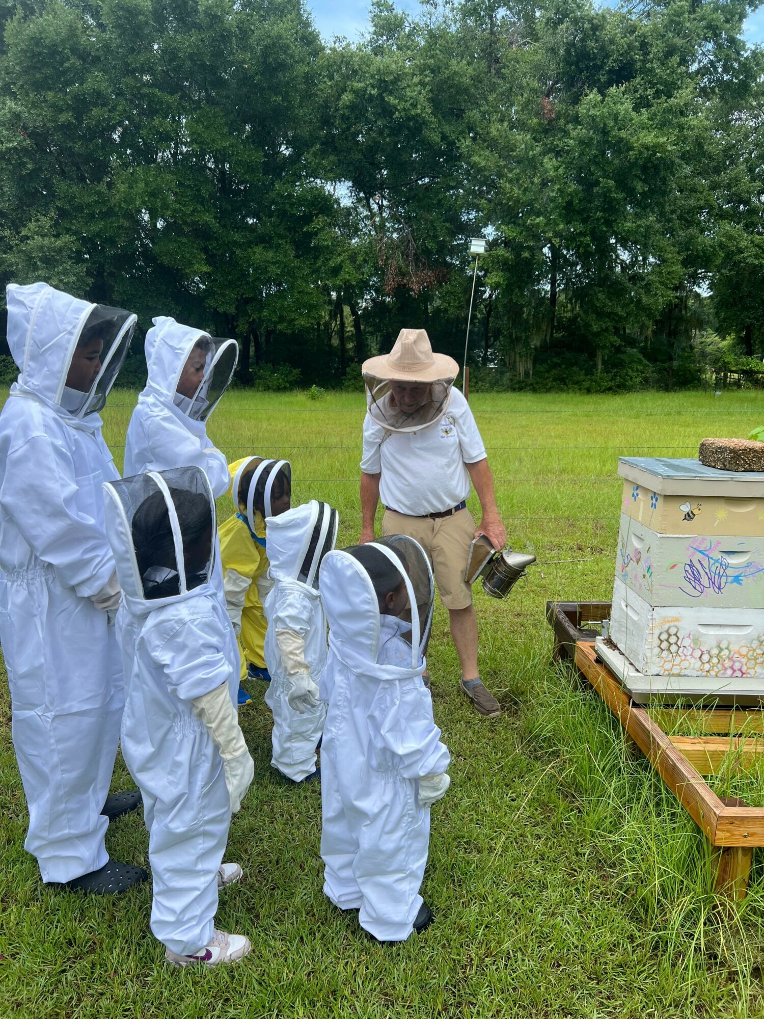 Person in hat and white shirt instructs children in white beekeeping suits outdoors.