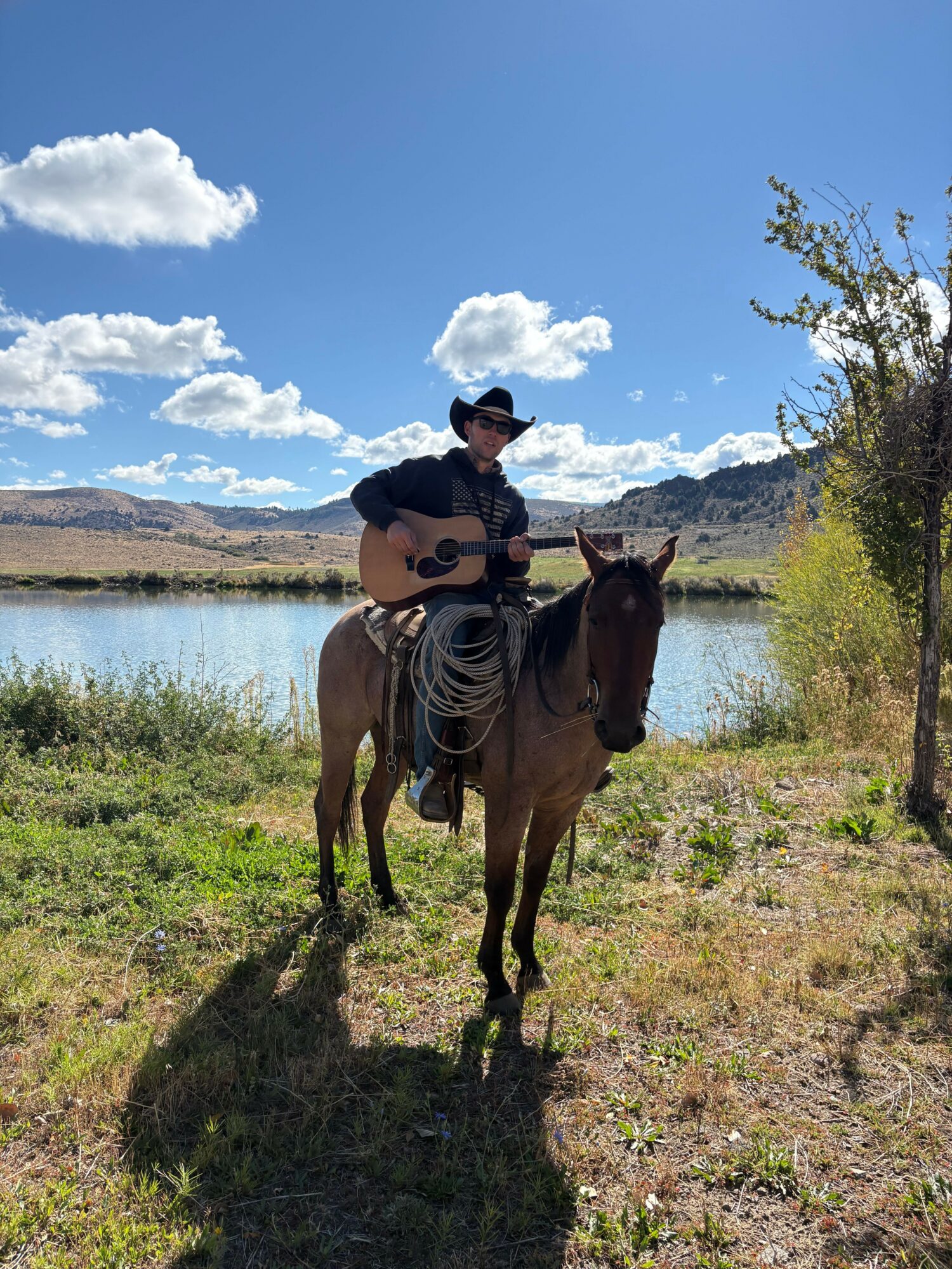 Person wearing a hat playing guitar while sitting on a horse near a lake with hills and trees in the background.
