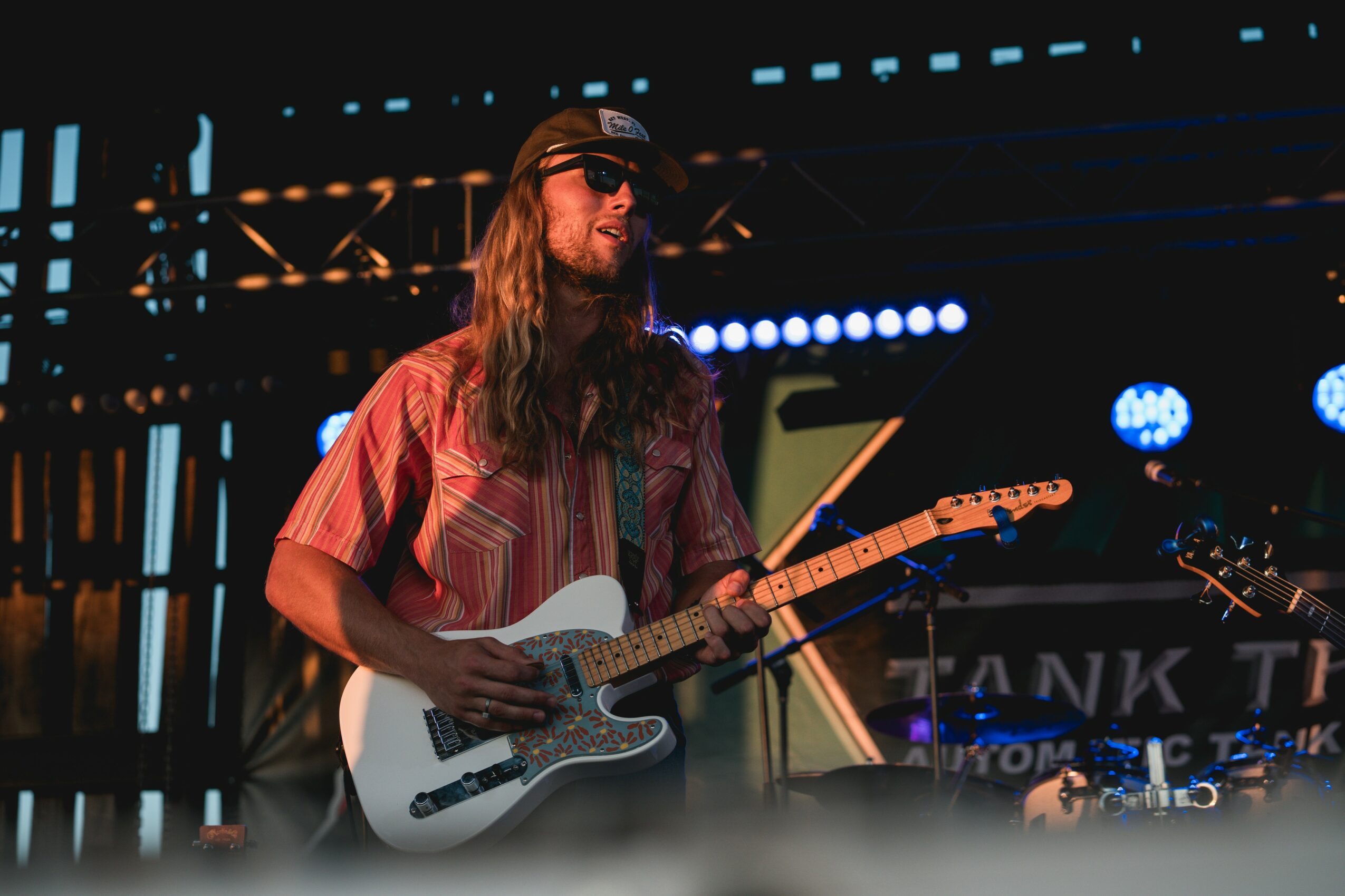 Musician with long hair, sunglasses, and a striped shirt playing an electric guitar on stage.