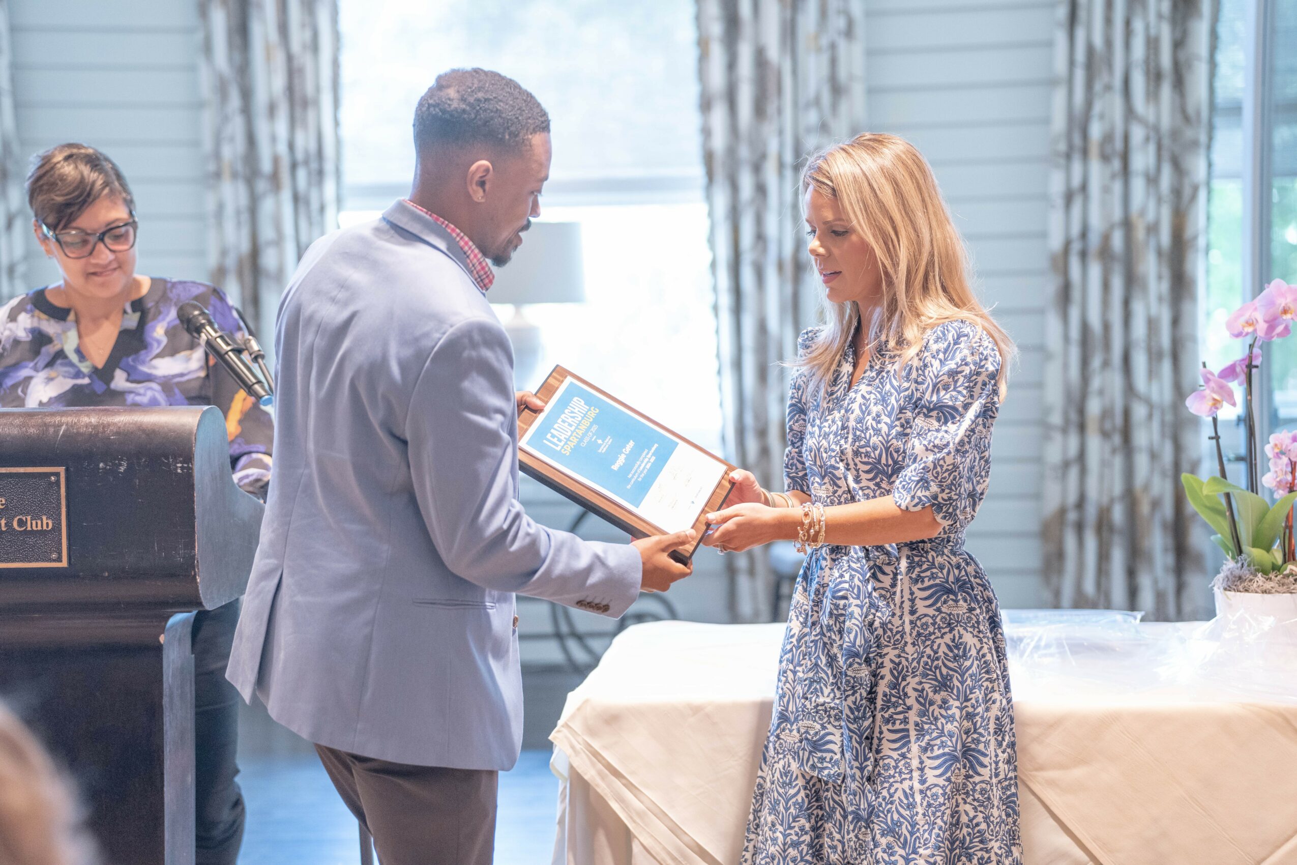 Man and woman exchanging a framed certificate in a room with a table and curtains.