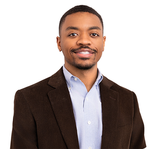 Man in a dark blazer and light blue shirt smiling against a white background.