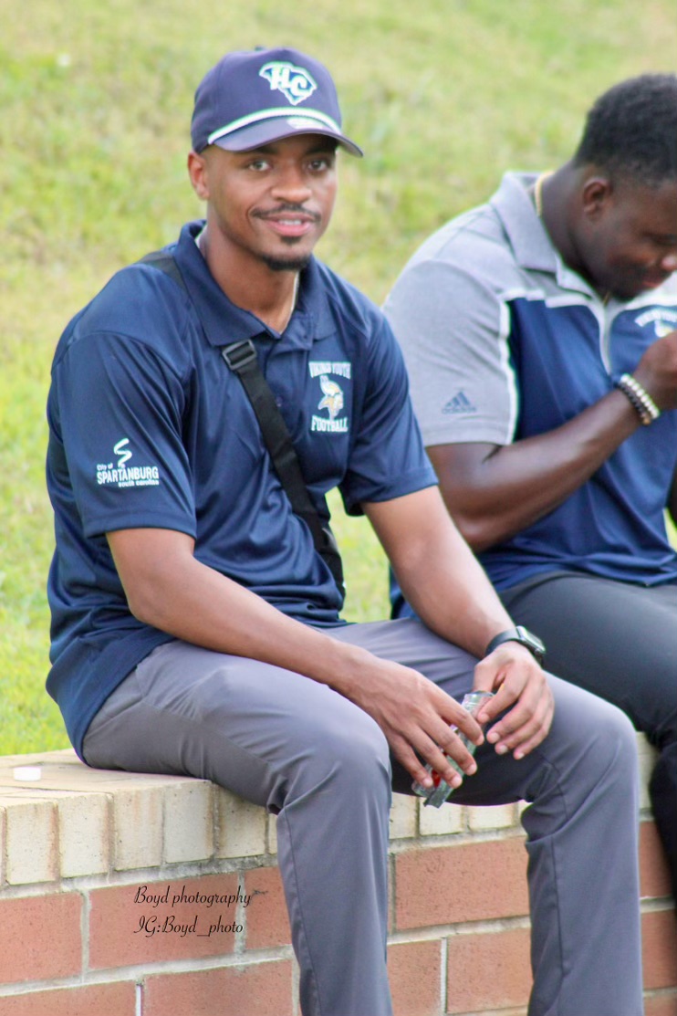 Man sitting outdoors on a brick ledge, smiling, wearing a navy blue cap and shirt, with another person beside him.