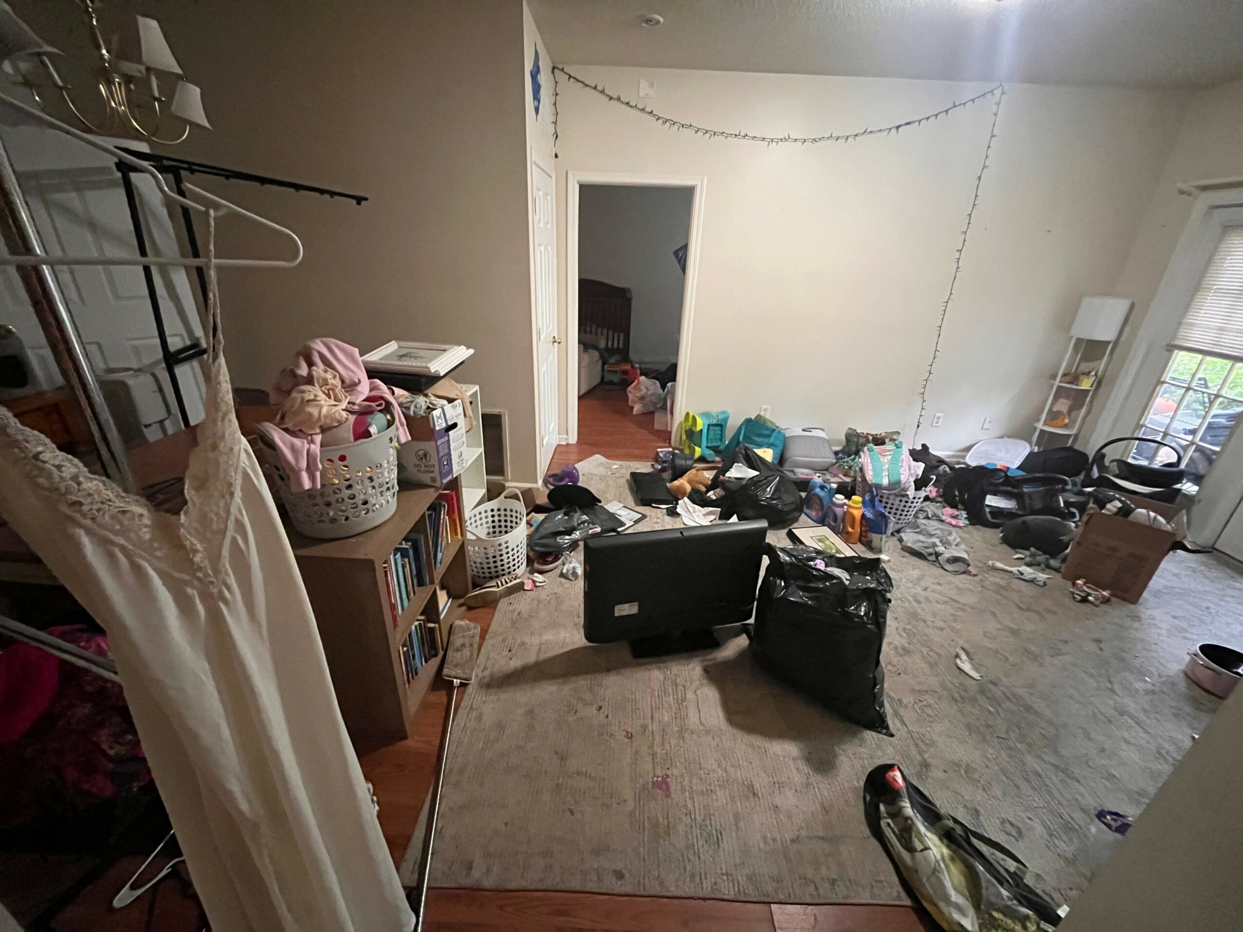 Room with cluttered floor, open laptop, bookshelf, and a doorway leading to another room, window with blinds on the right.