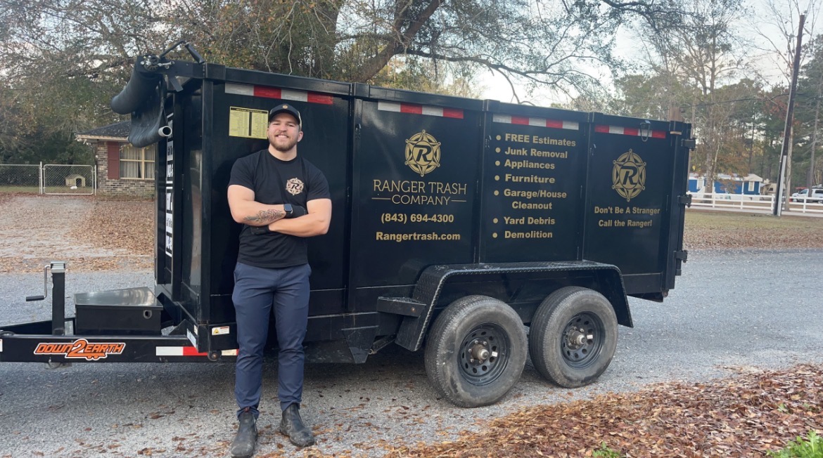 Person standing next to a black Ranger Trash vehicle outdoors with trees and a parking lot in background.