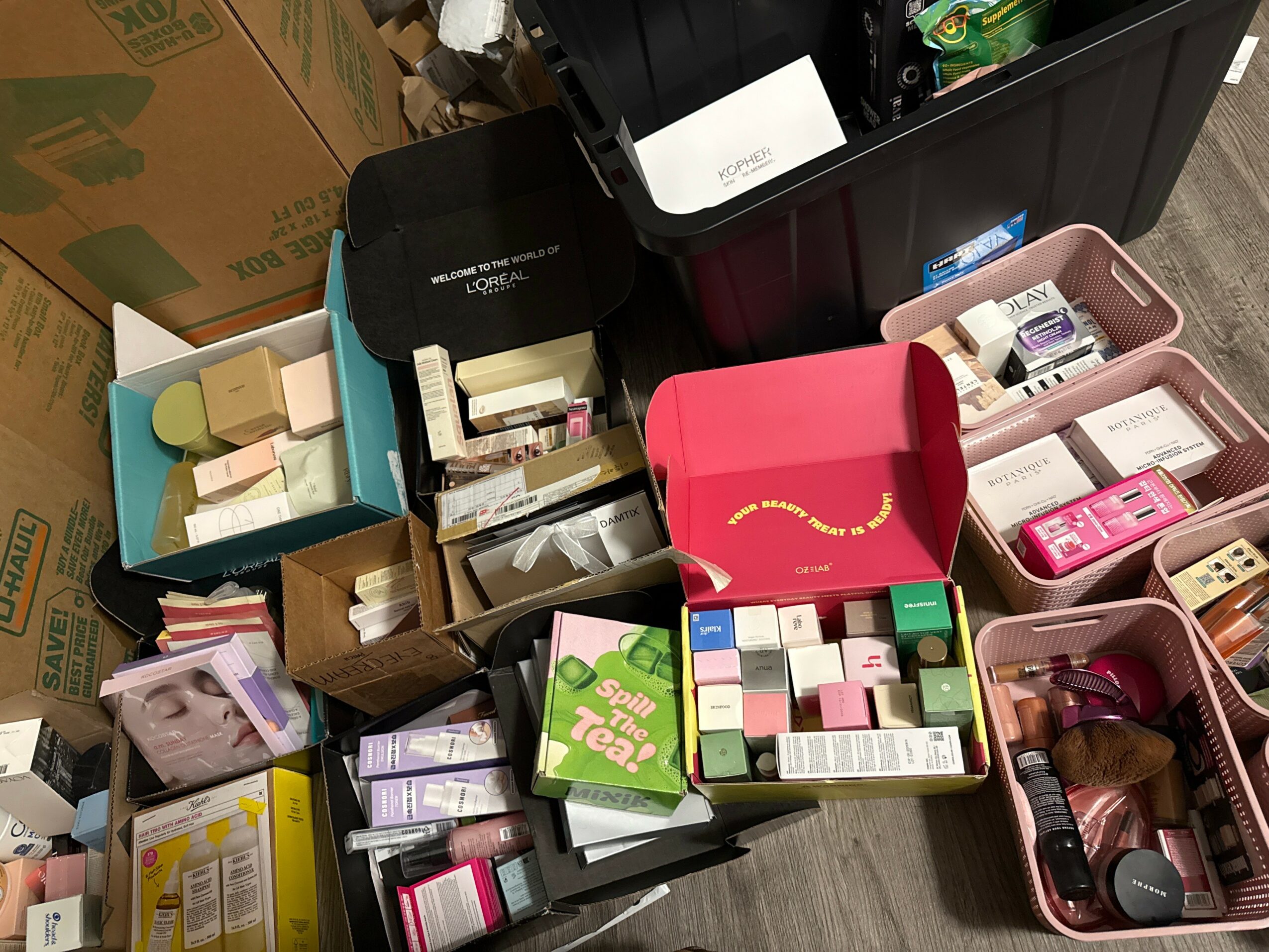 Open boxes and containers filled with various cosmetics and beauty products on a wooden floor.