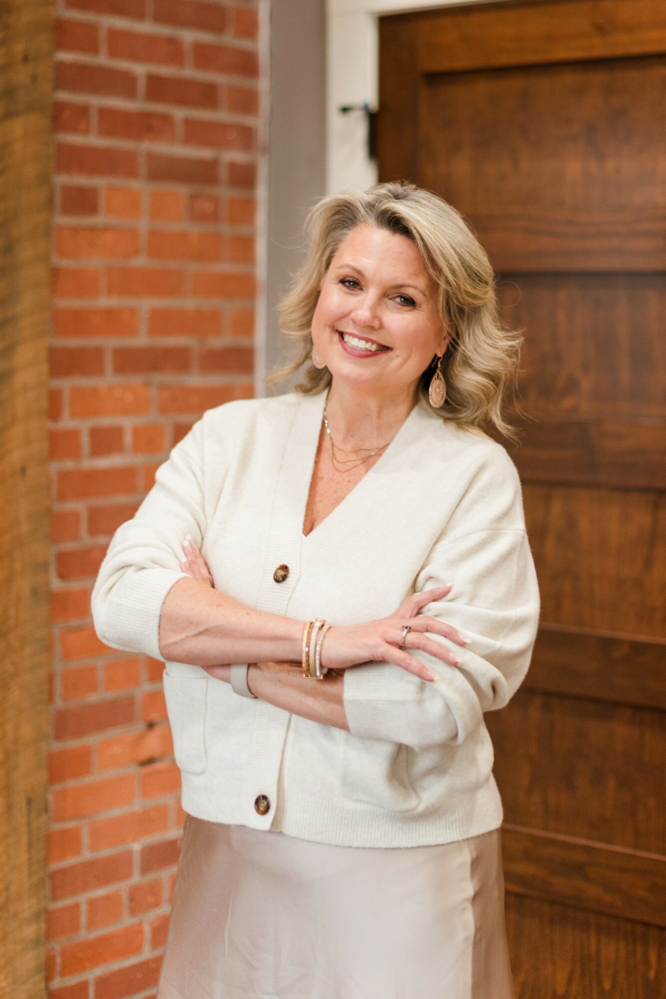 Smiling woman with blonde hair in a white jacket and skirt, standing outdoors near a brick wall and wooden door.