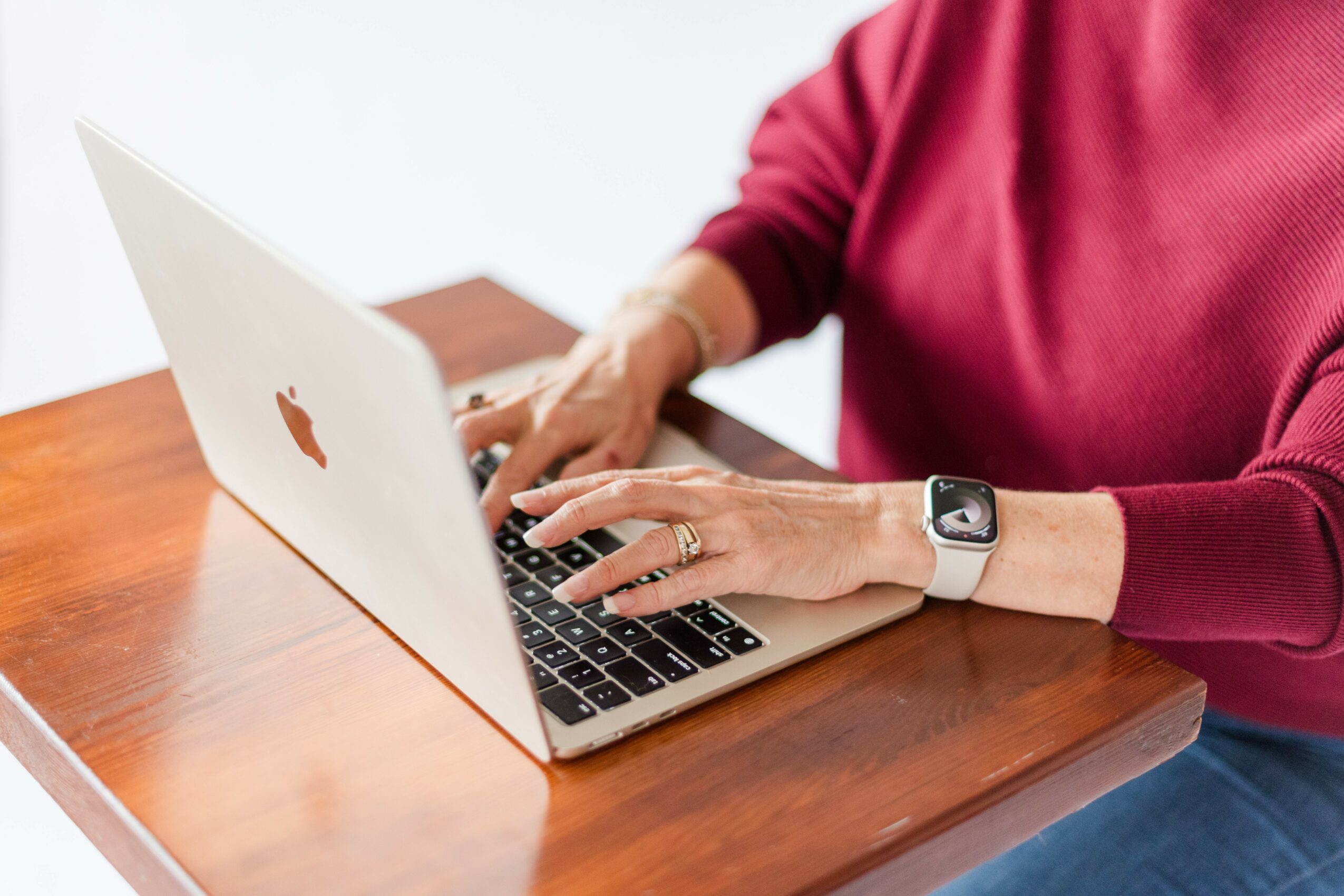 Person typing on a silver laptop with a wooden desk, wearing a red sweater and a watch.