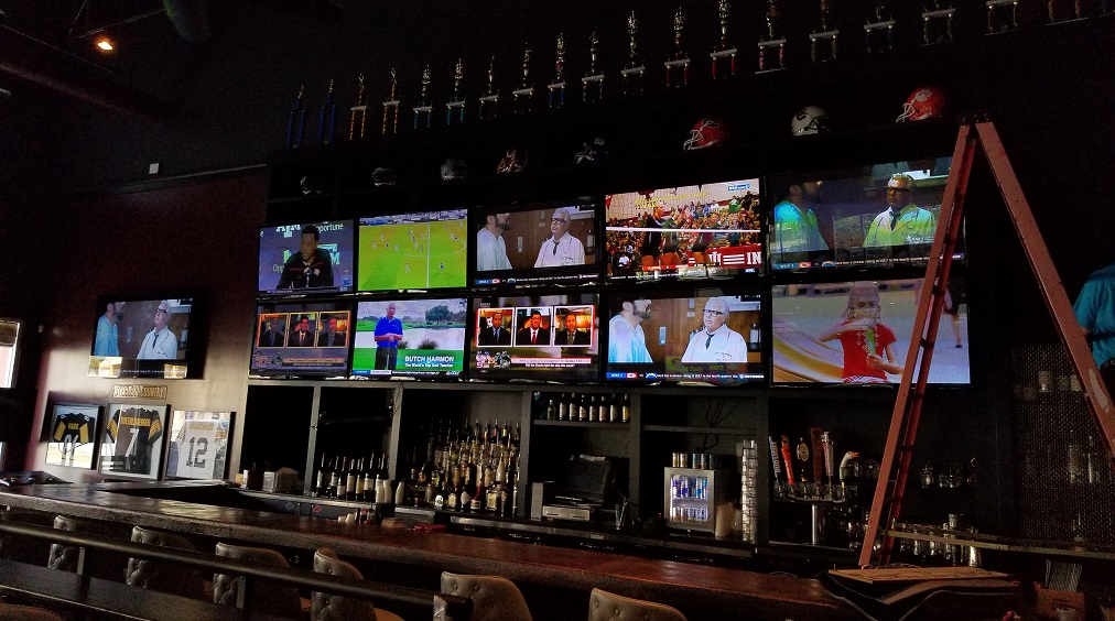 Multiple television screens displaying sports and news, bar shelves with bottles, and a ladder in a dimly lit bar.