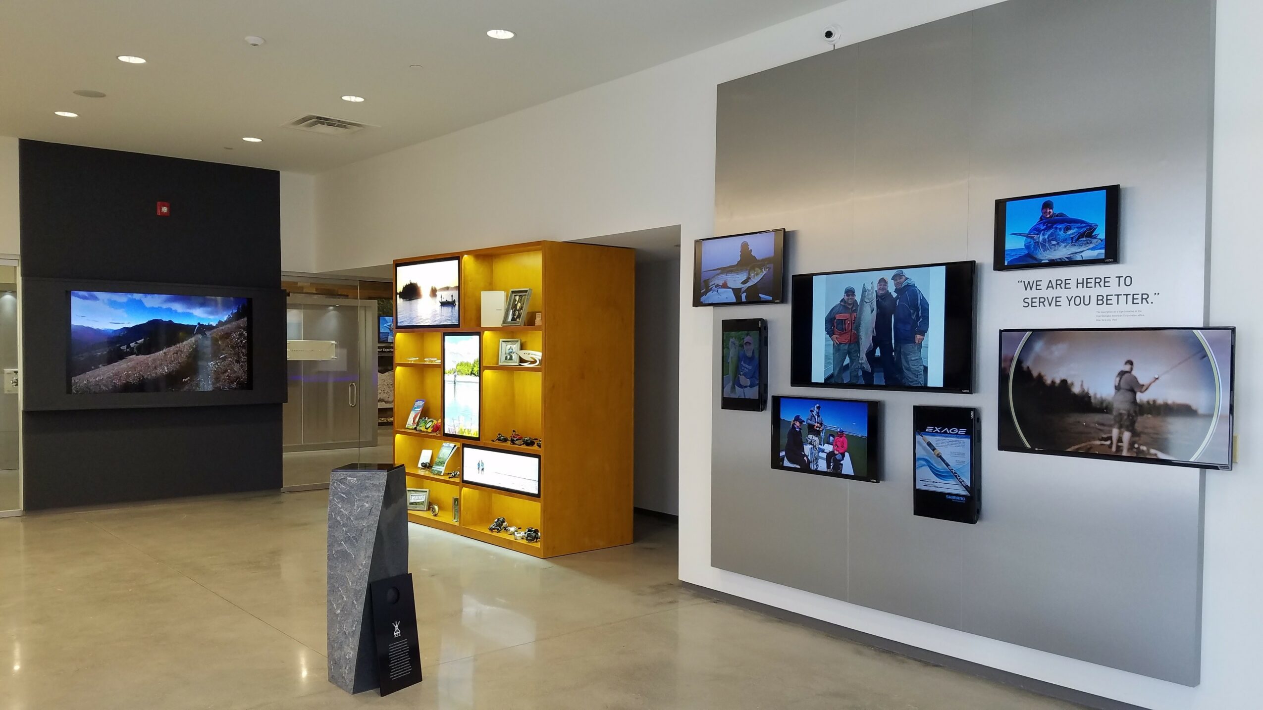 Gallery with wall-mounted screens displaying various images and a yellow display shelf with objects inside.