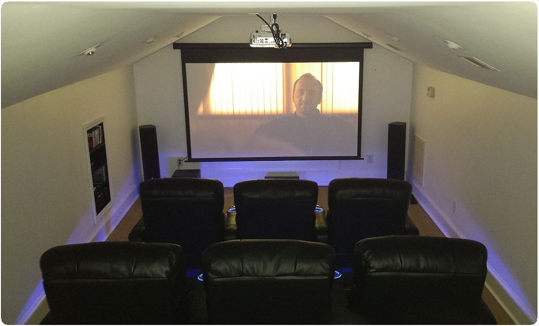 Home theater room with six black chairs facing a large screen and speakers, ceiling-mounted projector, and purple accent lighting.