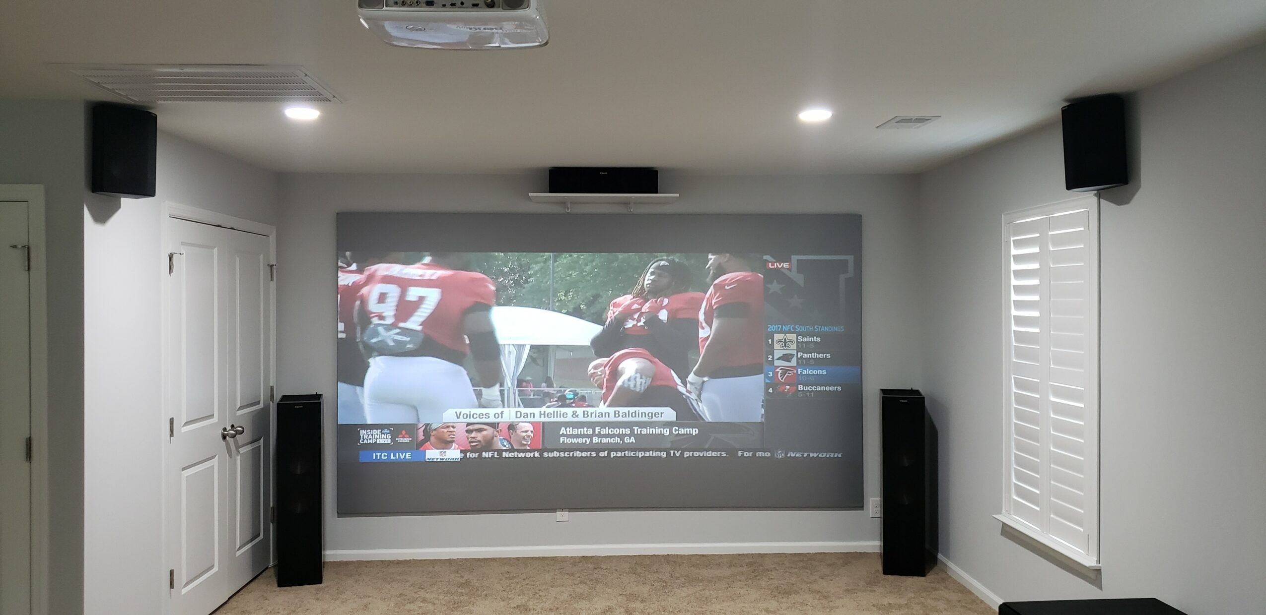 Home theater room with a large screen showing a football game, surrounded by speakers and white walls.