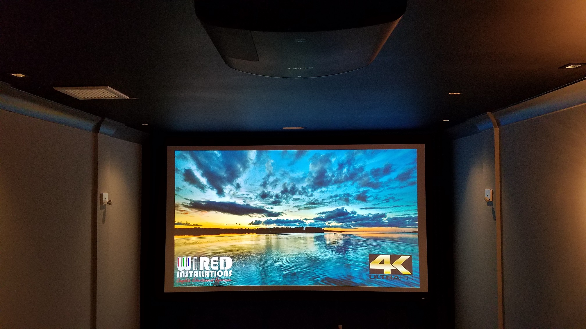 Projection screen displaying a landscape with water and sky, in a dark room with walls and ceiling visible.