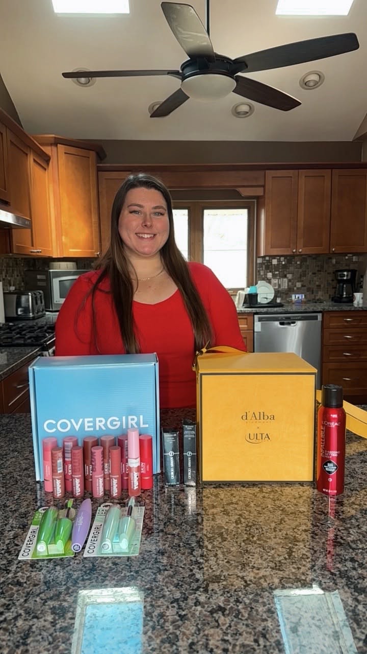 Woman standing behind a kitchen counter with makeup products and boxes, smiling at the camera.