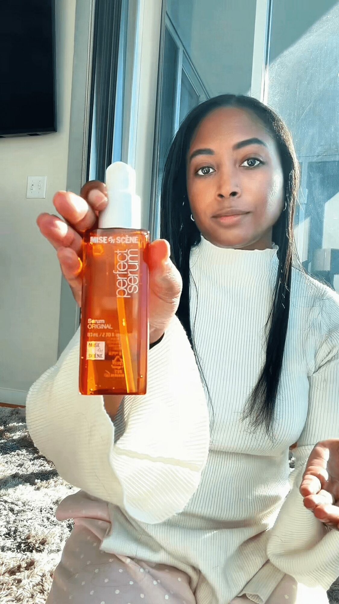 Young woman holding a bottle of orange skincare product indoors near window.