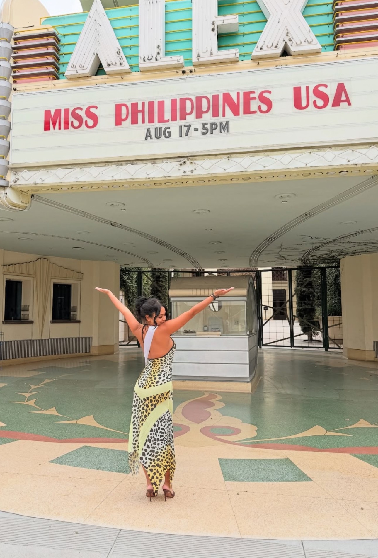 Person standing with arms raised in front of a theater marquee displaying Miss Philippines USA, August 17, 5 PM.