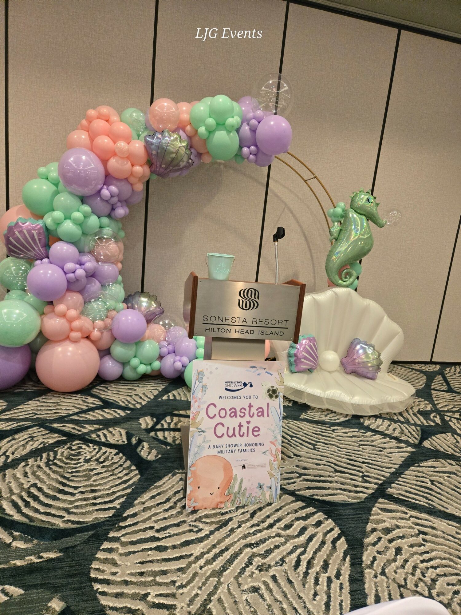 Decorative balloon arch with pastel colors, a seahorse figure, and a white bench with purple accents on a patterned tablecloth.