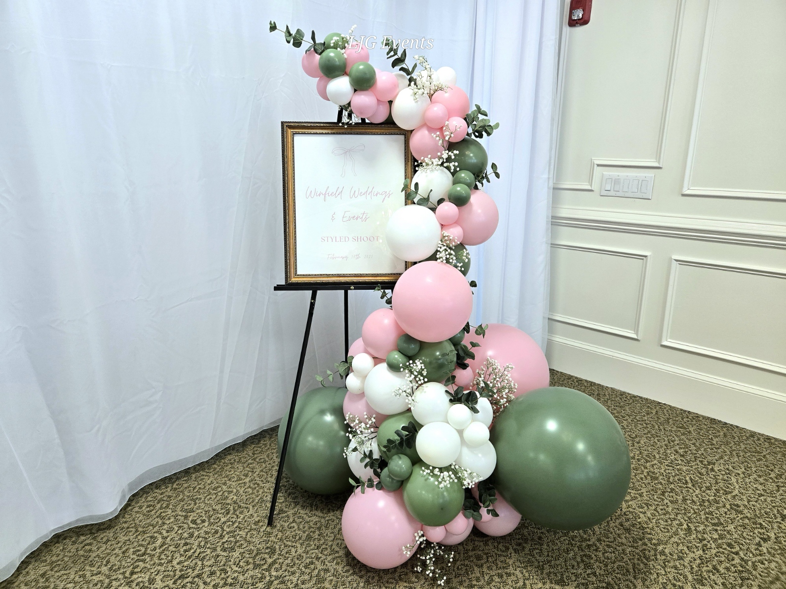Decorative balloon arrangement with pink, white, and green balloons and greenery, next to a framed sign on an easel.