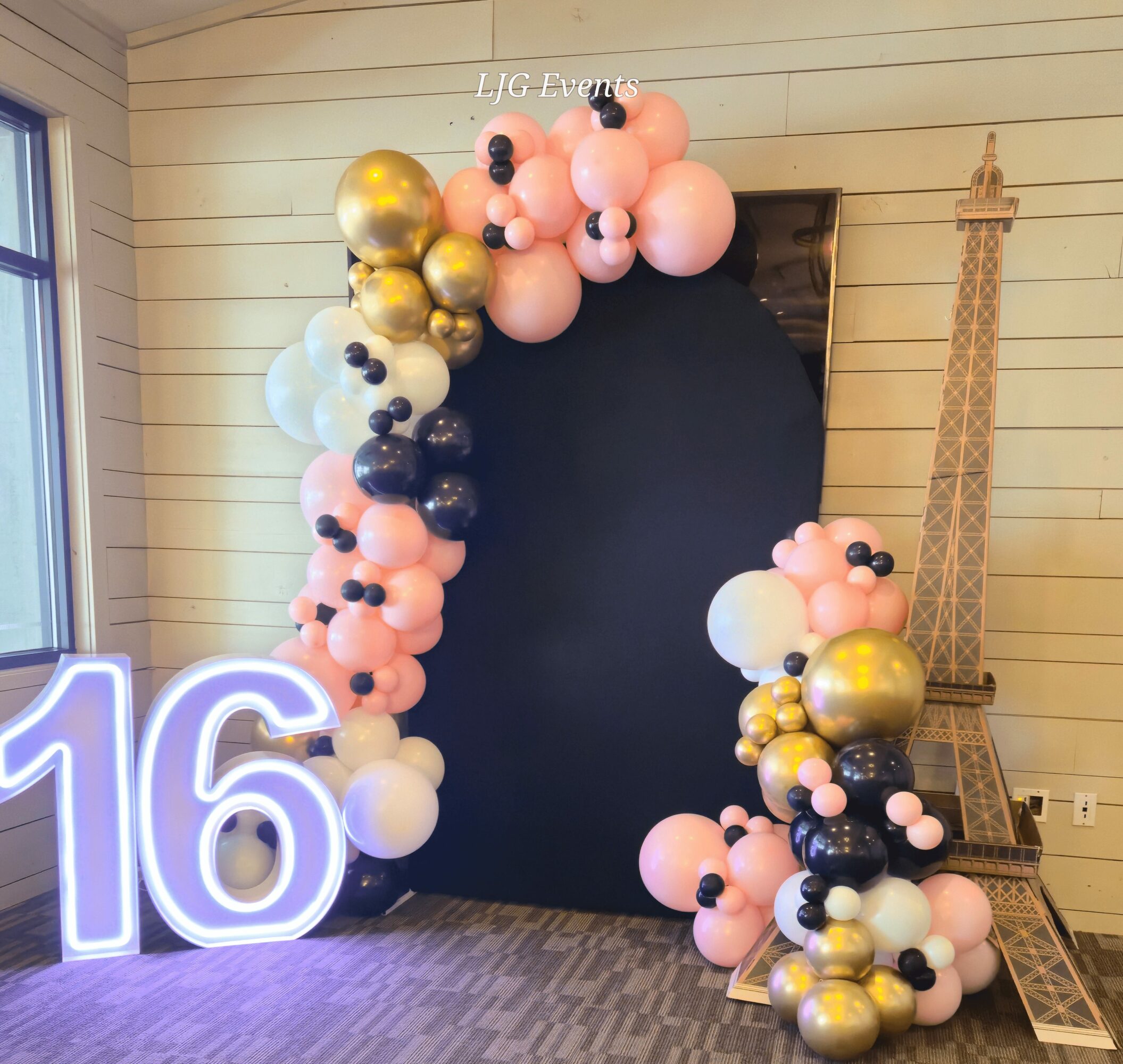 Balloon arch with pink, white, black, and gold balloons surrounds a black backdrop, with a large illuminated number 16 and a model of the Eiffel Tower nearby.