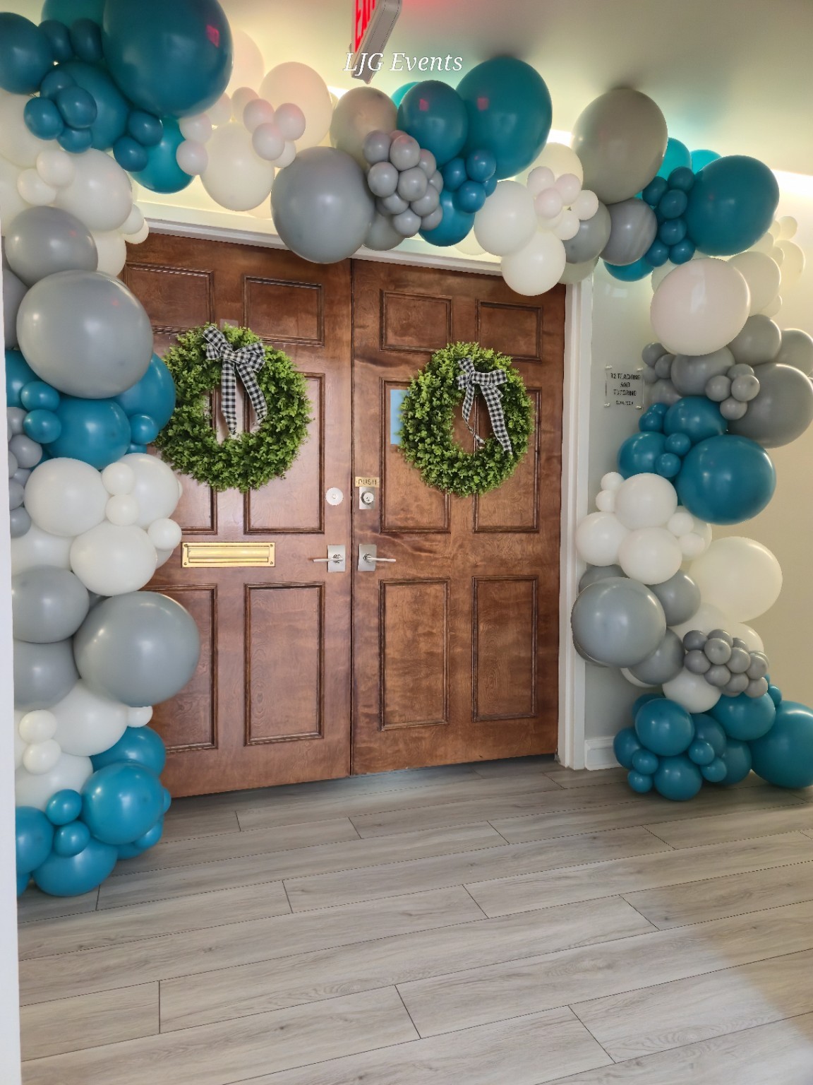 Double wooden door with wreaths, surrounded by balloon arch in blue, white, gray, and pink colors, on a wooden floor.