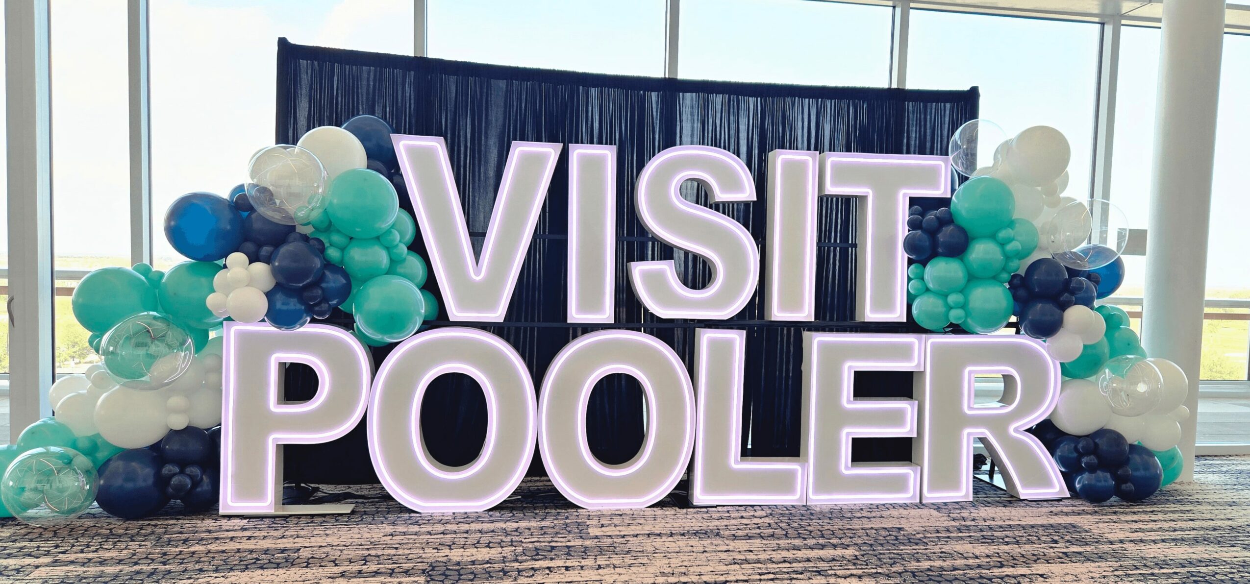 Large illuminated sign reads 'VISIT POOLER' with balloon decorations in front, inside a building with windows.
