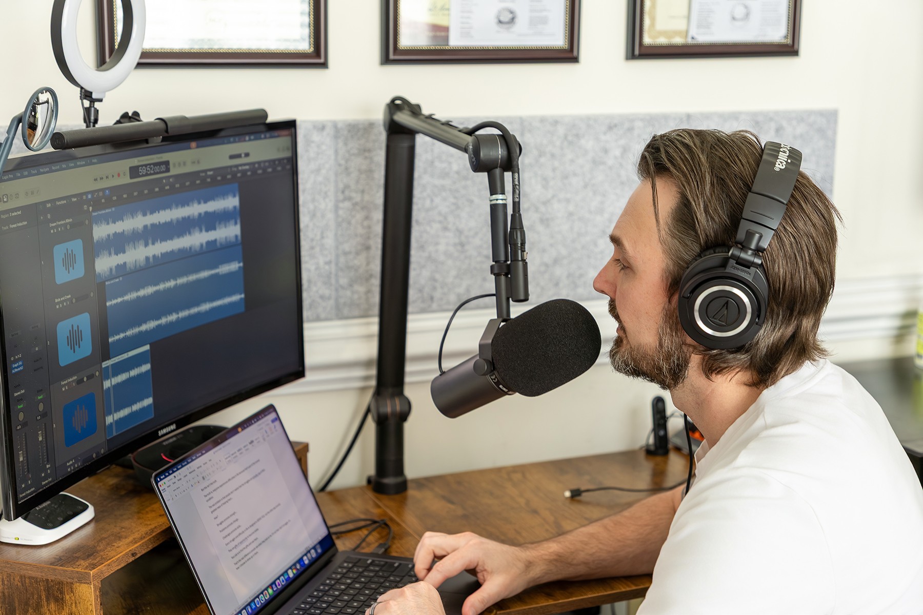 Man with headphones speaking into a microphone in a recording studio, with computer screens and framed certificates on the wall.