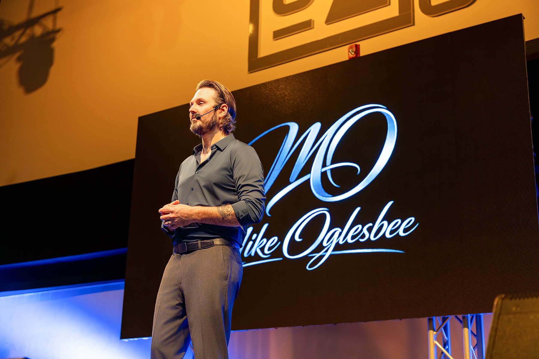 Man standing on stage with a large screen displaying 'MO like Oglesbee' behind him.