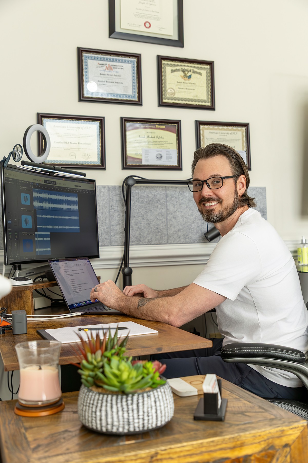 Man with glasses and beard working at desk with computer, certificates on wall behind him, plant and drink on desk.