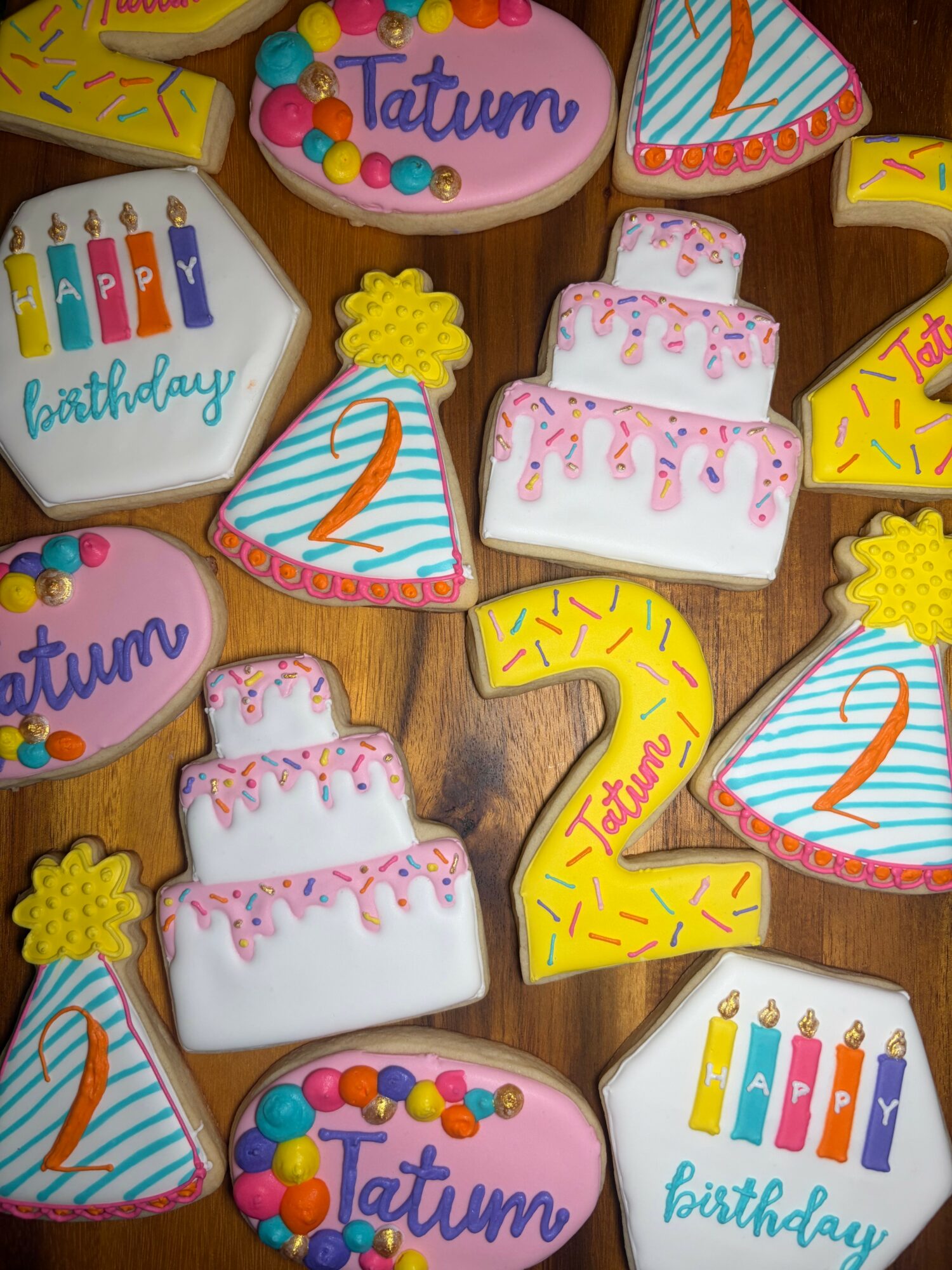 Assorted decorated cookies with birthday themes, including cakes, candles, and the name Tatum, on a wooden surface.